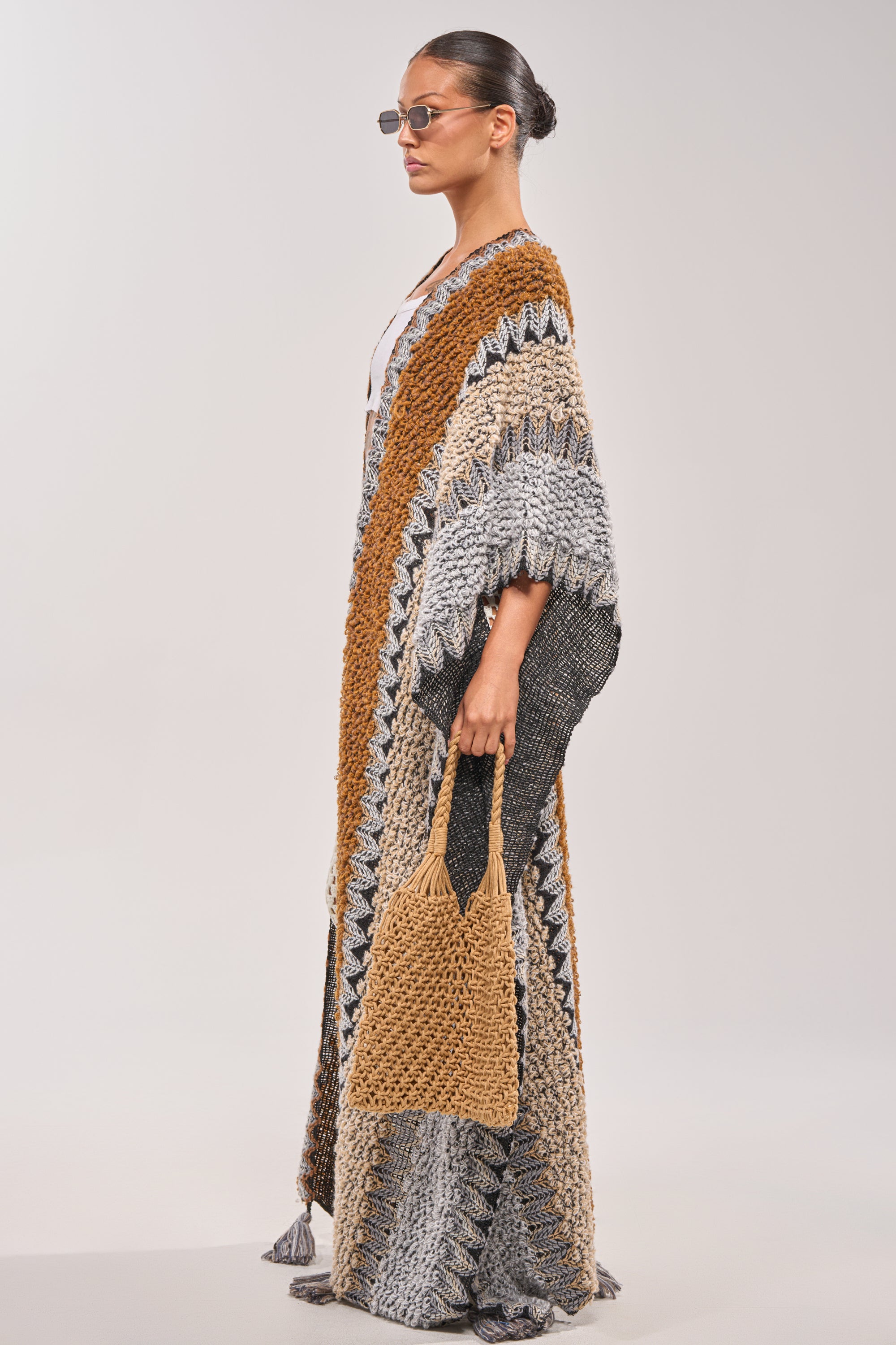 A woman in profile, wearing sunglasses and the CRAZY FOR YOU CARDIGAN in earth tones, holds a matching crocheted bag against a plain light background. 2