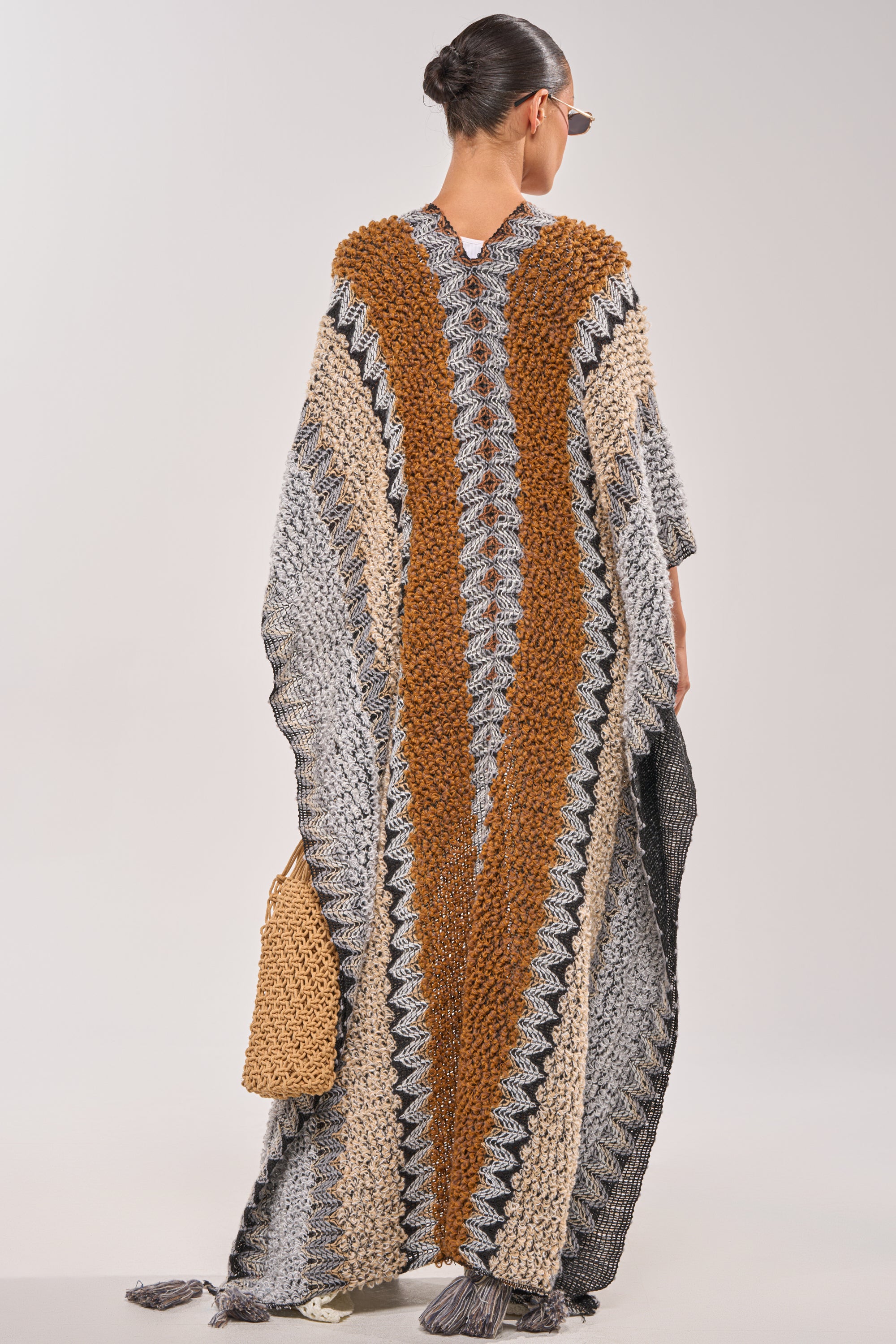 A woman with her hair in a bun and sunglasses stands facing away, holding a woven tan bag while wearing the CRAZY FOR YOU CARDIGAN—a long, striped piece with a robe-like silhouette in earth tones. 3