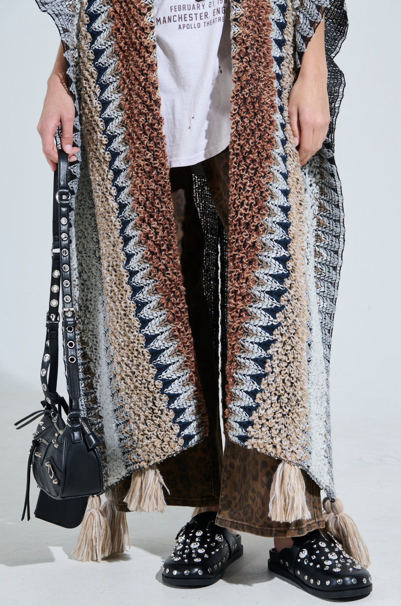 A person showcases a hippie-chic look, adorned in the CRAZY FOR YOU CARDIGAN in multicolored crochet with tassels over a white graphic T-shirt. They accessorize with a black studded handbag, brown patterned pants, and black studded shoes to complete the bohemian ensemble.