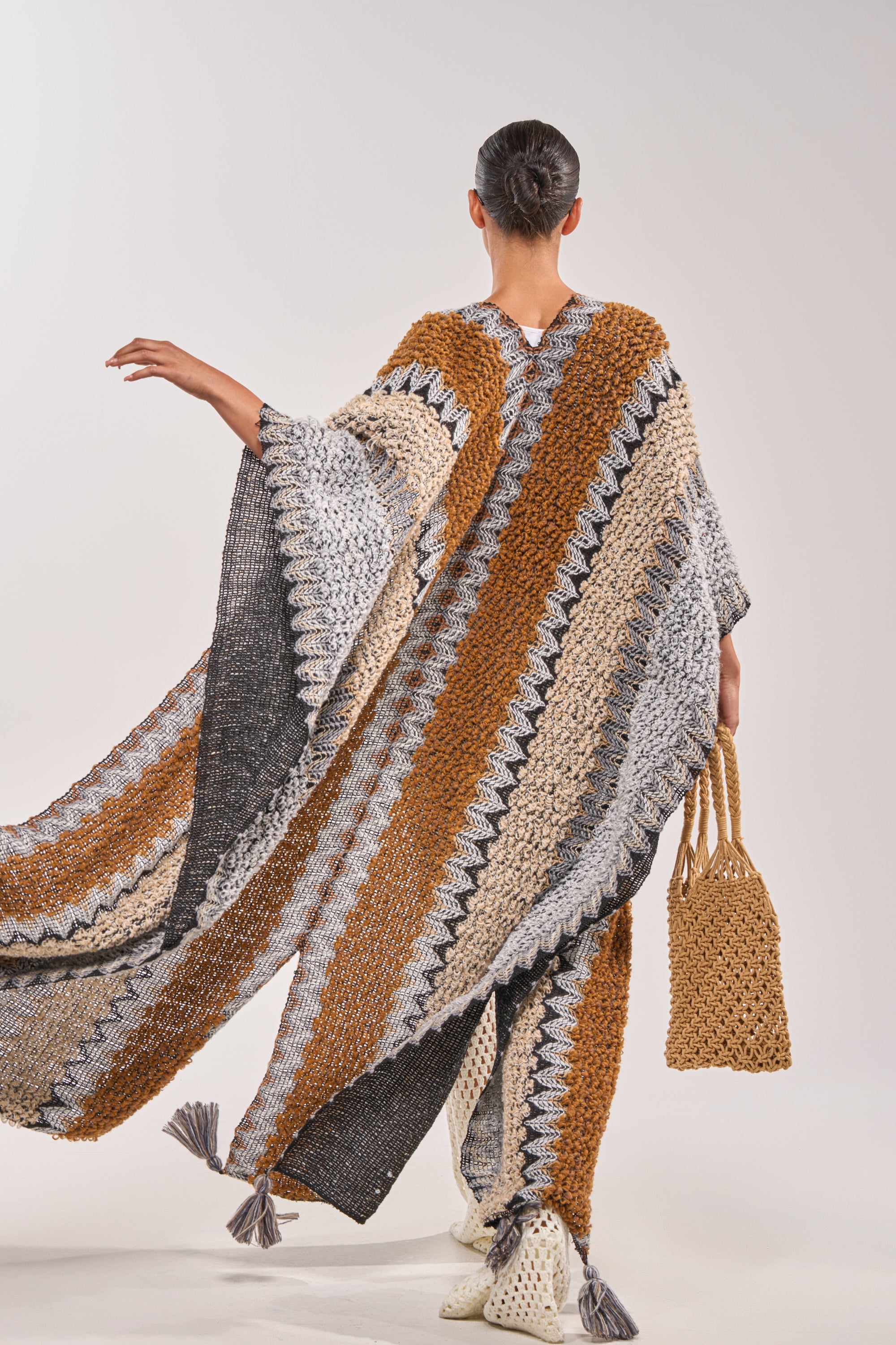 A person stands facing away, wearing the CRAZY FOR YOU CARDIGAN—a long, wide-sleeved striped piece in brown, gray, black, and cream. They carry a woven bag, wear white shoes, and have their hair neatly tied back. 4