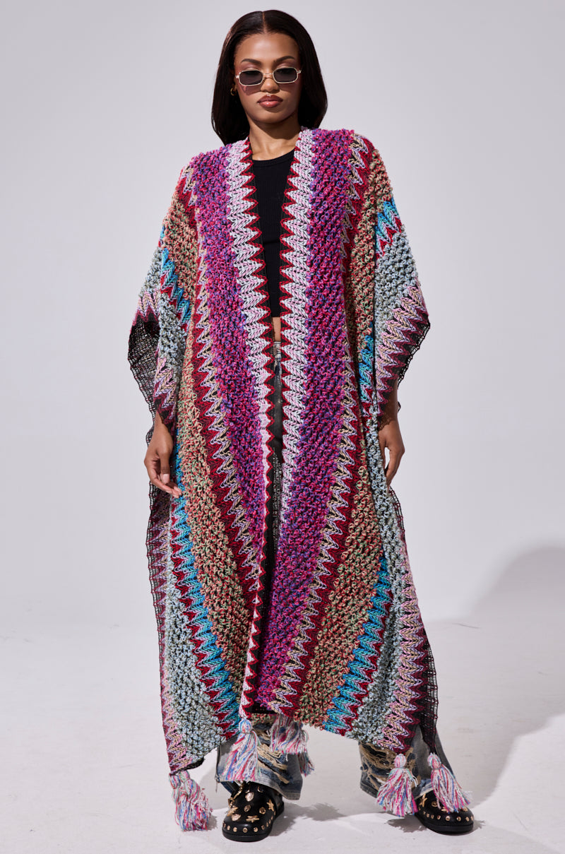 A woman wearing sunglasses and the FALL IN LOVE WITH ME CARDIGAN, featuring pink, blue, and beige zigzag patterns, stands confidently against a plain background. She completes her look with black studded shoes.