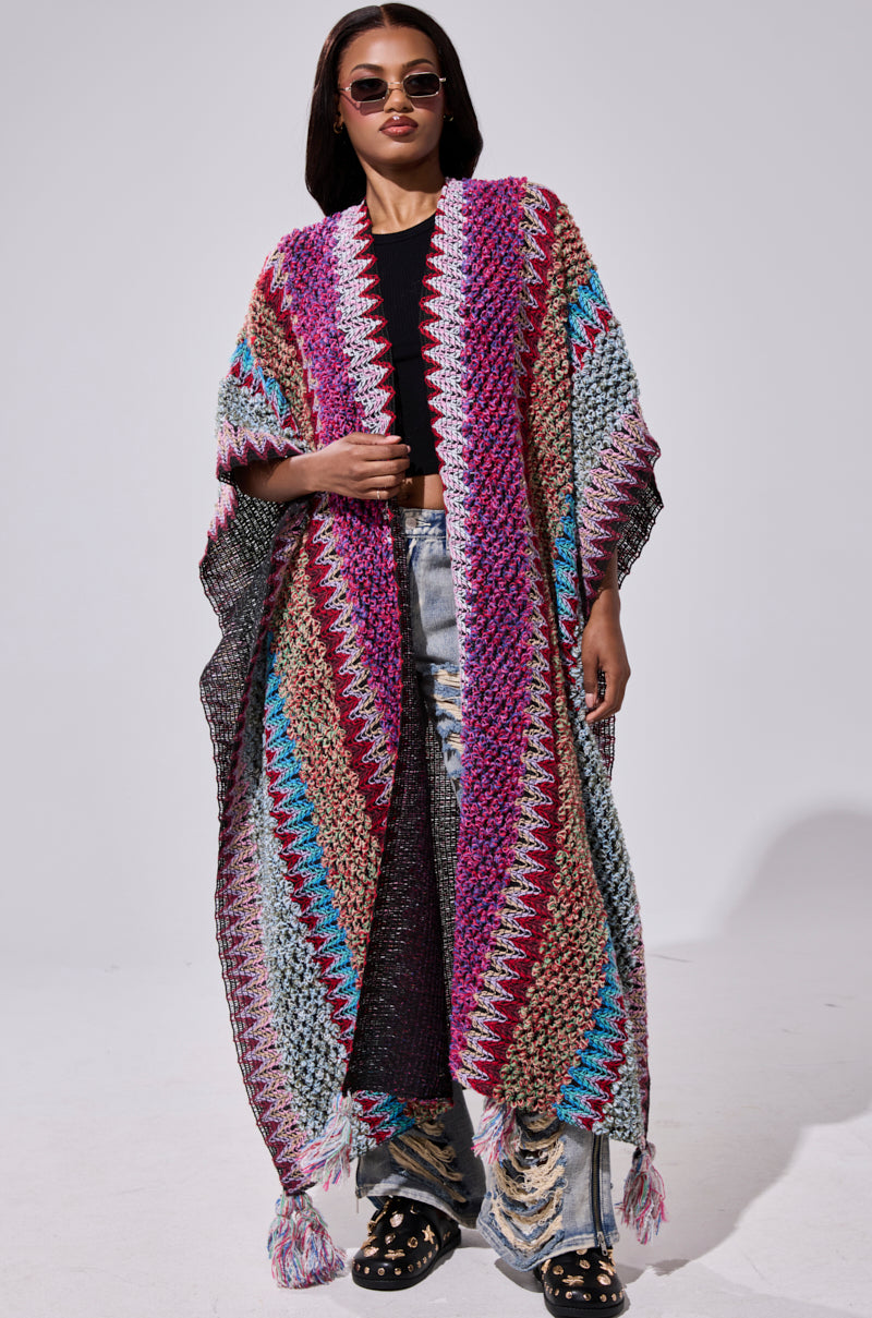 A woman confidently poses against a plain background wearing sunglasses, a black top, distressed jeans, studded black shoes, and the FALL IN LOVE WITH ME CARDIGAN—a colorful, oversized crocheted sweater with a zigzag pattern.