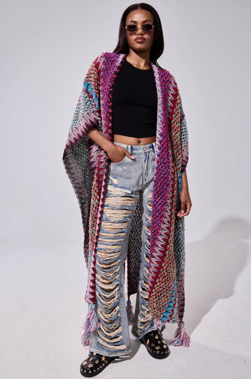 A woman stands confidently against a plain background wearing sunglasses, a black crop top, light blue distressed jeans, star-patterned black shoes, and the FALL IN LOVE WITH ME CARDIGAN—a colorful crochet piece with tassels.