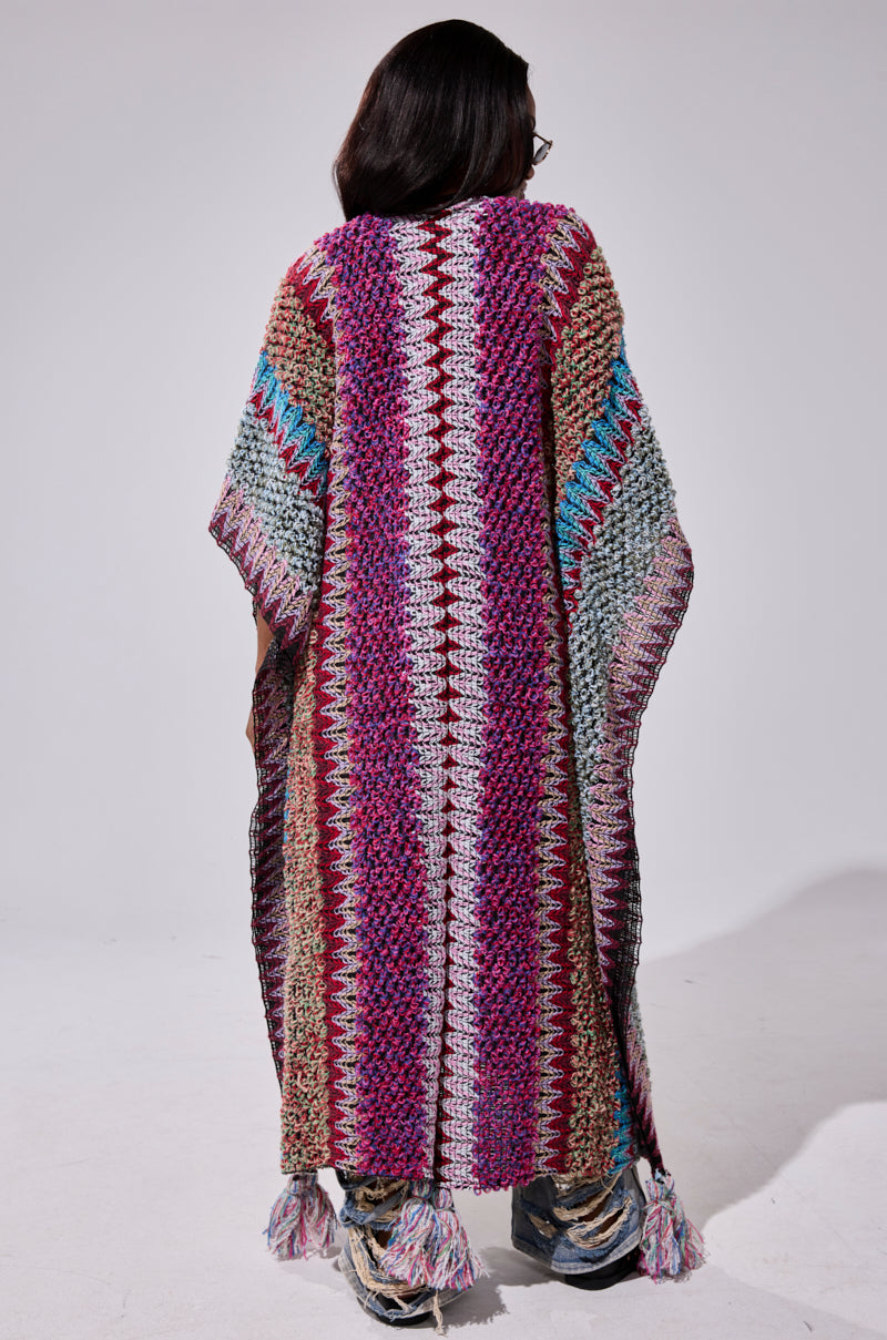 A person with long dark hair, seen from behind, wears the oversized FALL IN LOVE WITH ME CARDIGAN—a vibrant, striped knit poncho with tassels in shades of purple, red, green, blue, and cream.