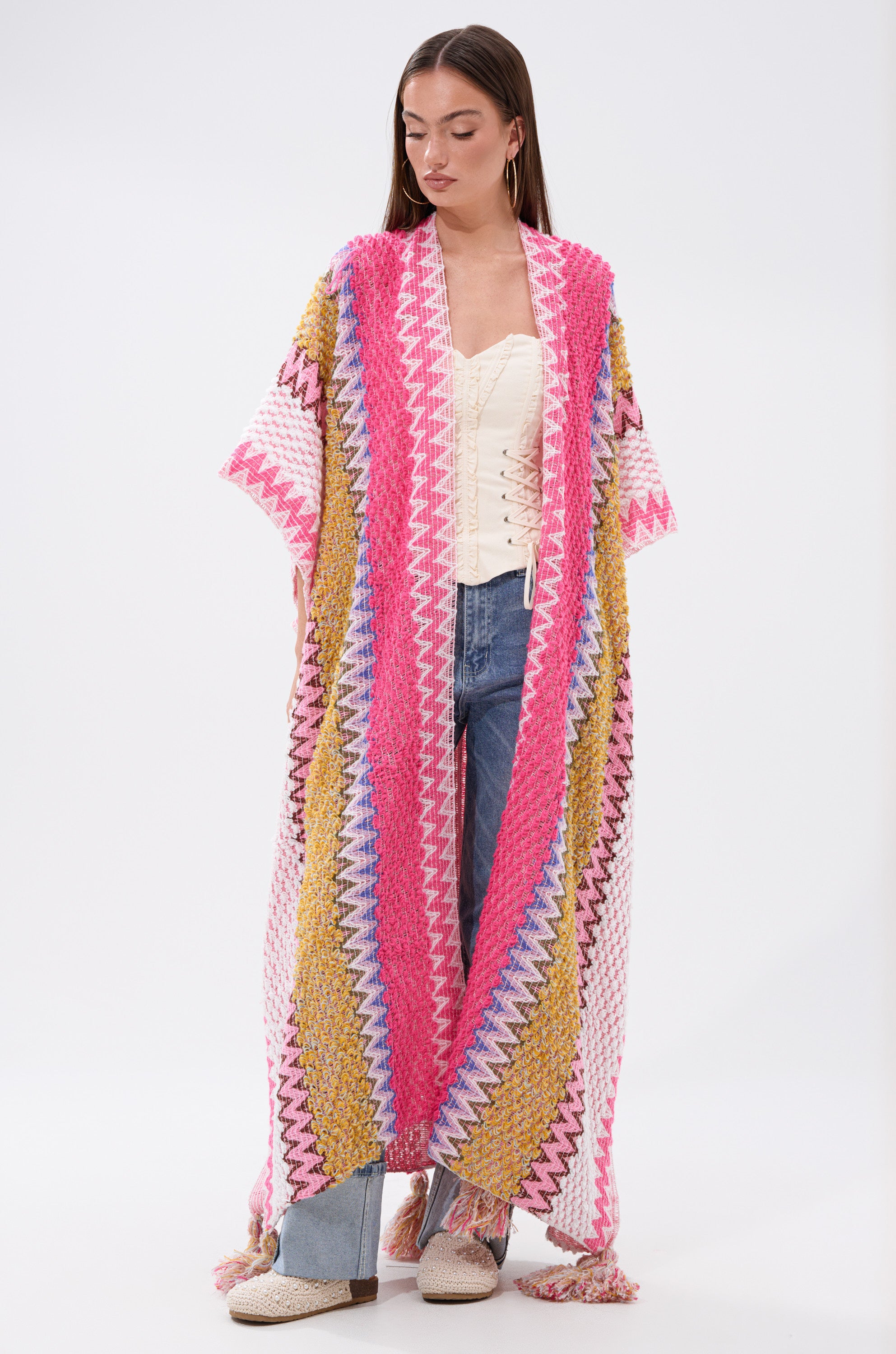 A woman models the DREAM STRIPED CARDIGAN—an oversized knit with pink, yellow, white, and blue chevron stripes—layered over a white top and light blue jeans against a plain white background.
