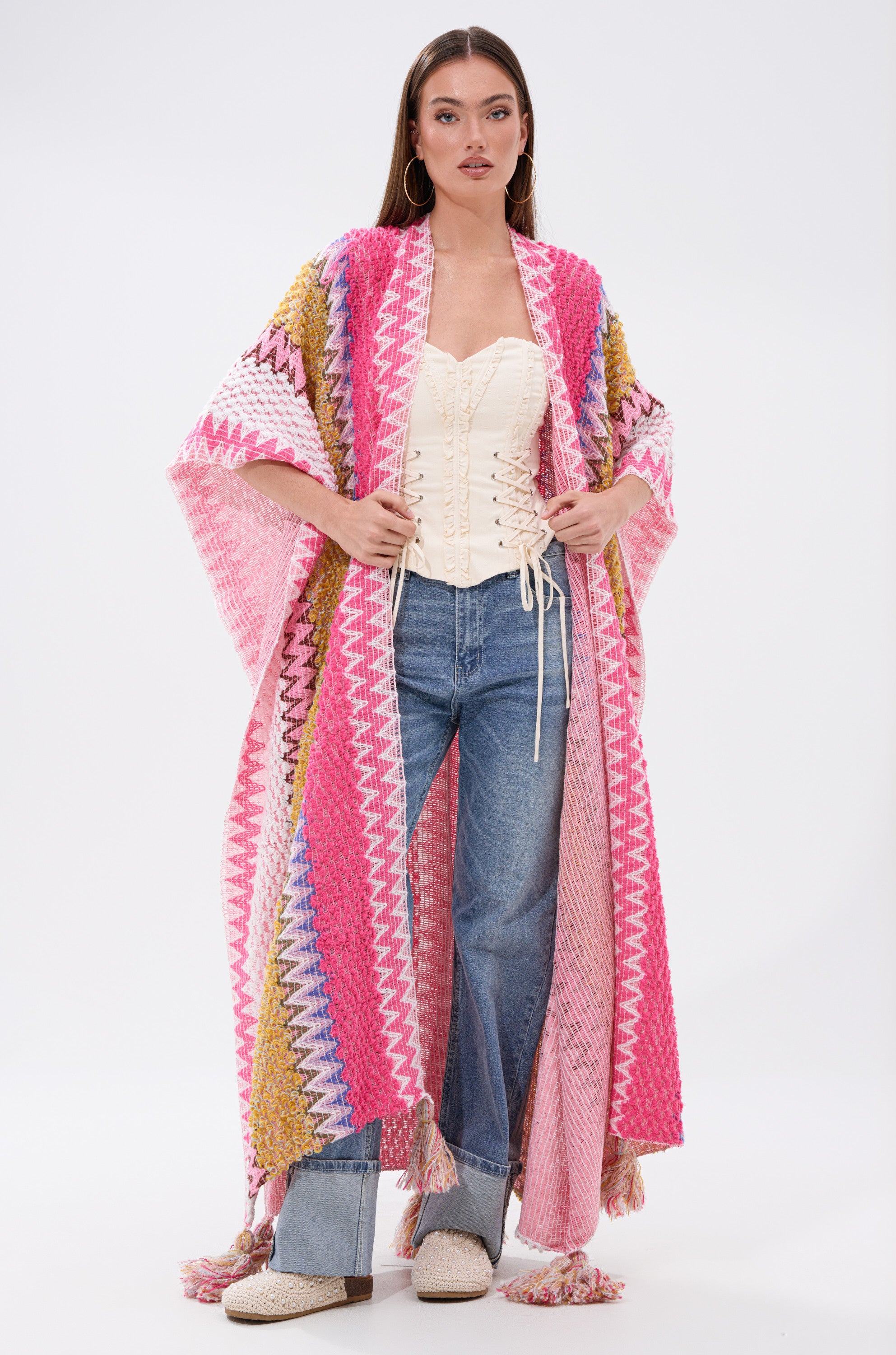 A woman wears the DREAM STRIPED CARDIGAN over a white corset and blue jeans.