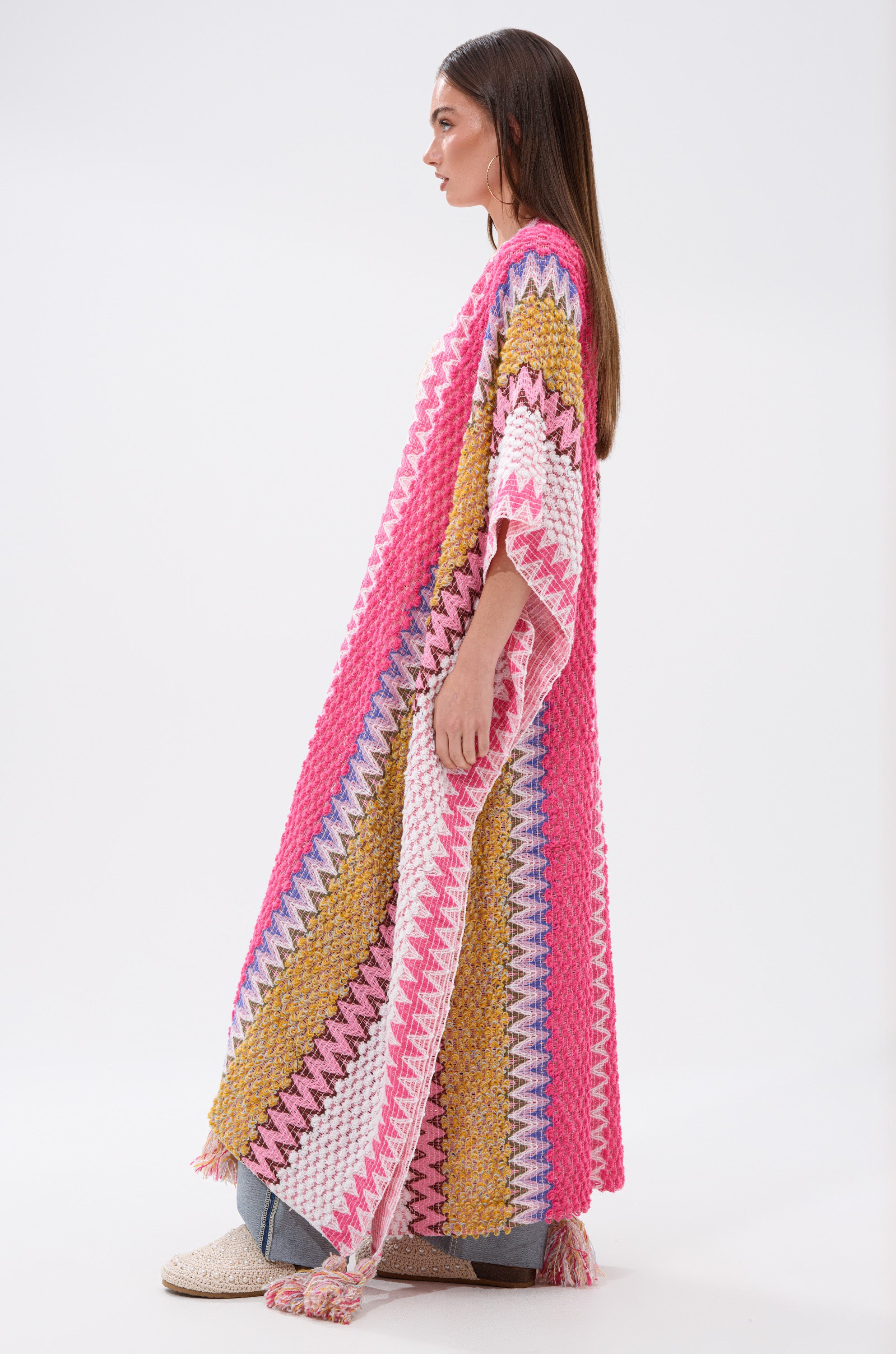 A woman in profile wears the DREAM STRIPED CARDIGAN—a long, colorful knit robe with pink, yellow, white, and purple stripes—paired with blue jeans and cream shoes against a plain white background.