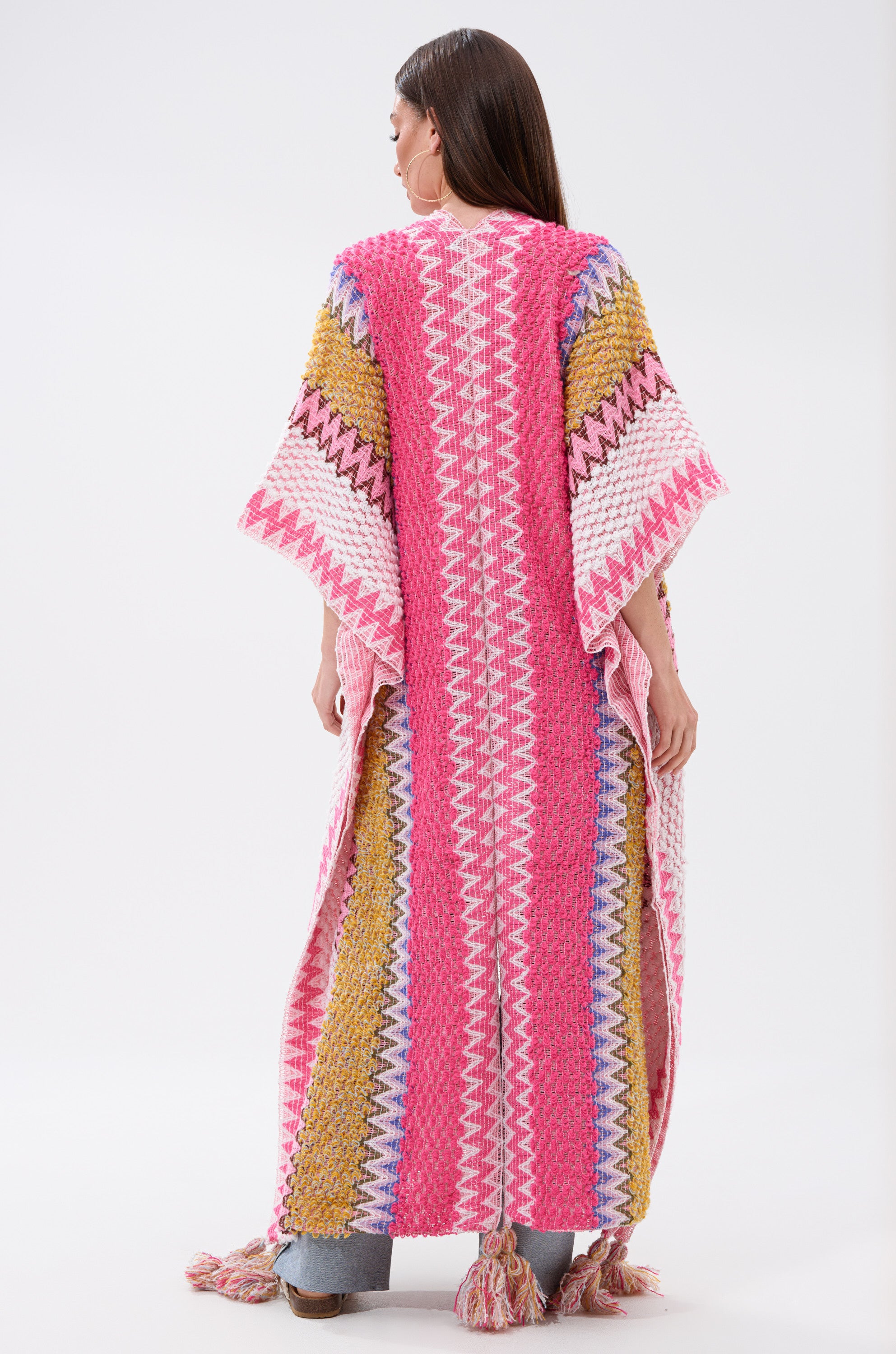 A woman faces away, wearing the DREAM STRIPED CARDIGAN—an oversized chevron knit with wide sleeves, tassels at the hem, and a bold zigzag pattern in pink, yellow, purple, white, and blue.