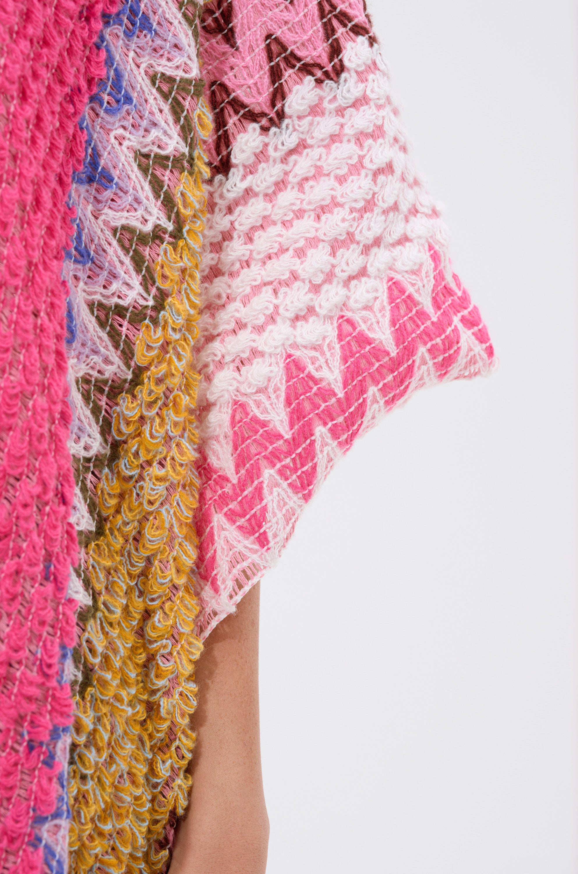 Close-up of the DREAM STRIPED CARDIGAN, showcasing its colorful chevron knit fabric in pink, white, yellow, brown, and purple zigzag patterns. A person's bare arm appears beside the cardigan against a plain white background.