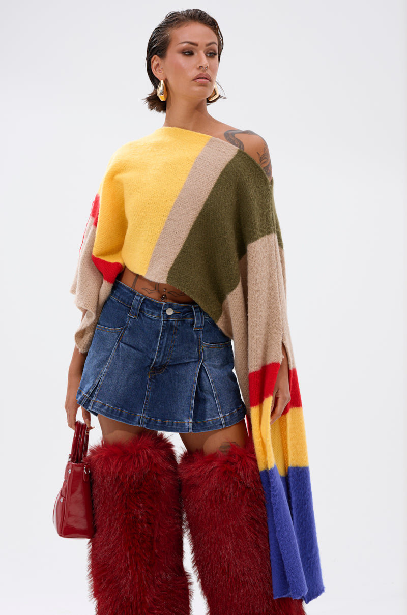 A woman wears the EASY LOVER CROP ABSTRACT SWEATER, a short denim skirt, tall red furry boots, gold hoop earrings, and holds a red handbag against a plain background.