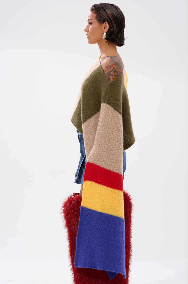 A woman with short brown hair and a large arm tattoo stands sideways, wearing the EASY LOVER CROP ABSTRACT SWEATER in olive green, beige, red, yellow, and blue stripes with denim shorts and holding a red furry bag.