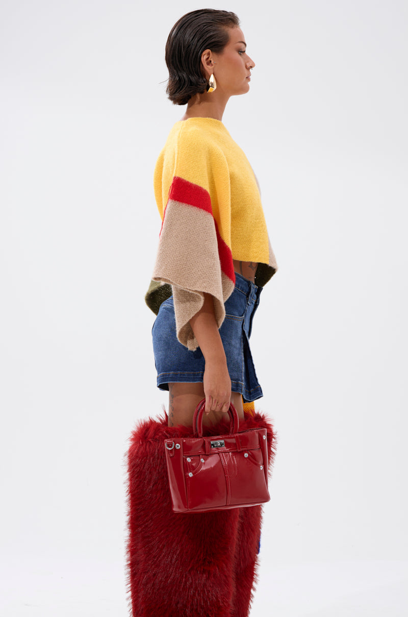 A woman in profile wears the EASY LOVER CROP ABSTRACT SWEATER in yellow, beige, and red with denim shorts and tall red furry boots. She carries a shiny red handbag and finishes her look with gold earrings, posed against a white background.