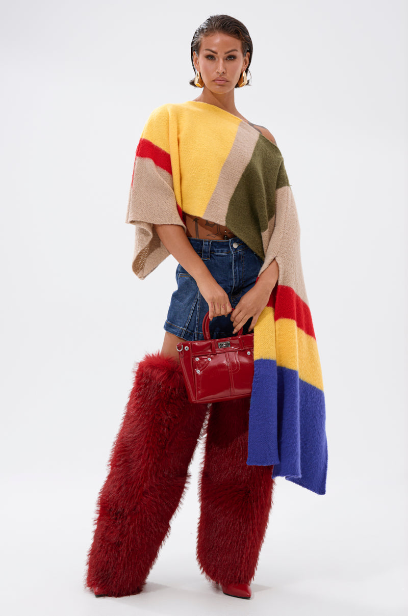 A woman stands against a plain background wearing the EASY LOVER CROP ABSTRACT SWEATER with denim shorts, artsy knit leg warmers, red heeled boots, and a small red handbag. She looks confidently at the camera.