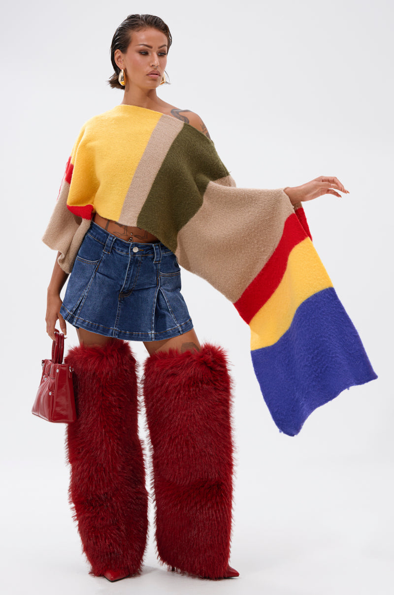 A woman wears the EASY LOVER CROP ABSTRACT SWEATER with a denim mini skirt and tall, furry red boots. She holds a small red handbag, her hair slicked back, standing against a plain white background.