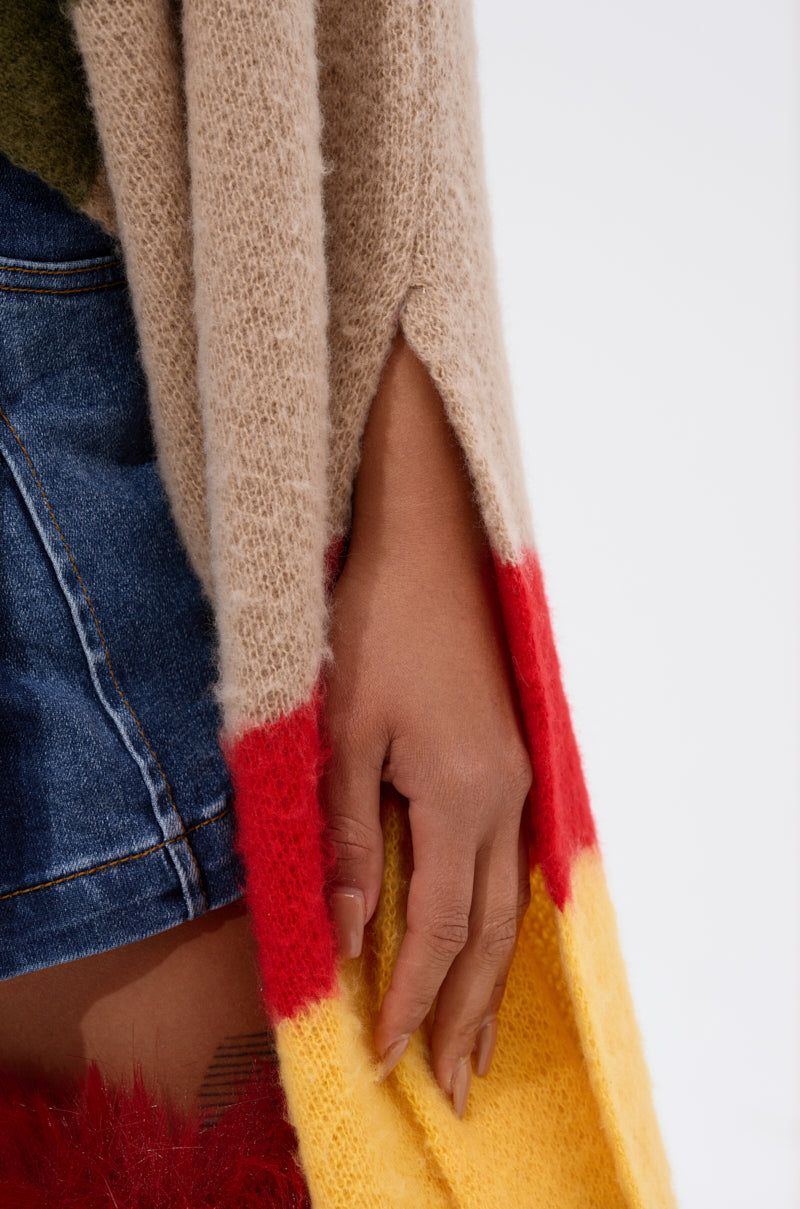 A close-up of a hand resting by the side, wearing the EASY LOVER CROP ABSTRACT SWEATER in beige, red, and yellow colorblock over blue denim shorts against a plain light background.