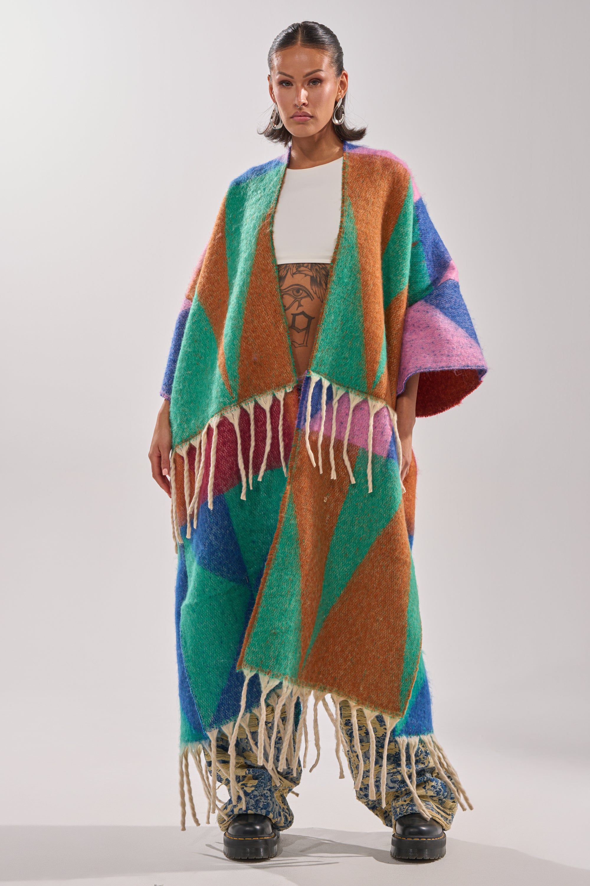 A woman exudes confidence in the FEELING CRAZY OVERSIZED CARDIGAN, a vibrant geometric fringed sweater styled over a white top and printed pants with chunky boots. Her hair is slicked back; the plain light background highlights her bold winter look.