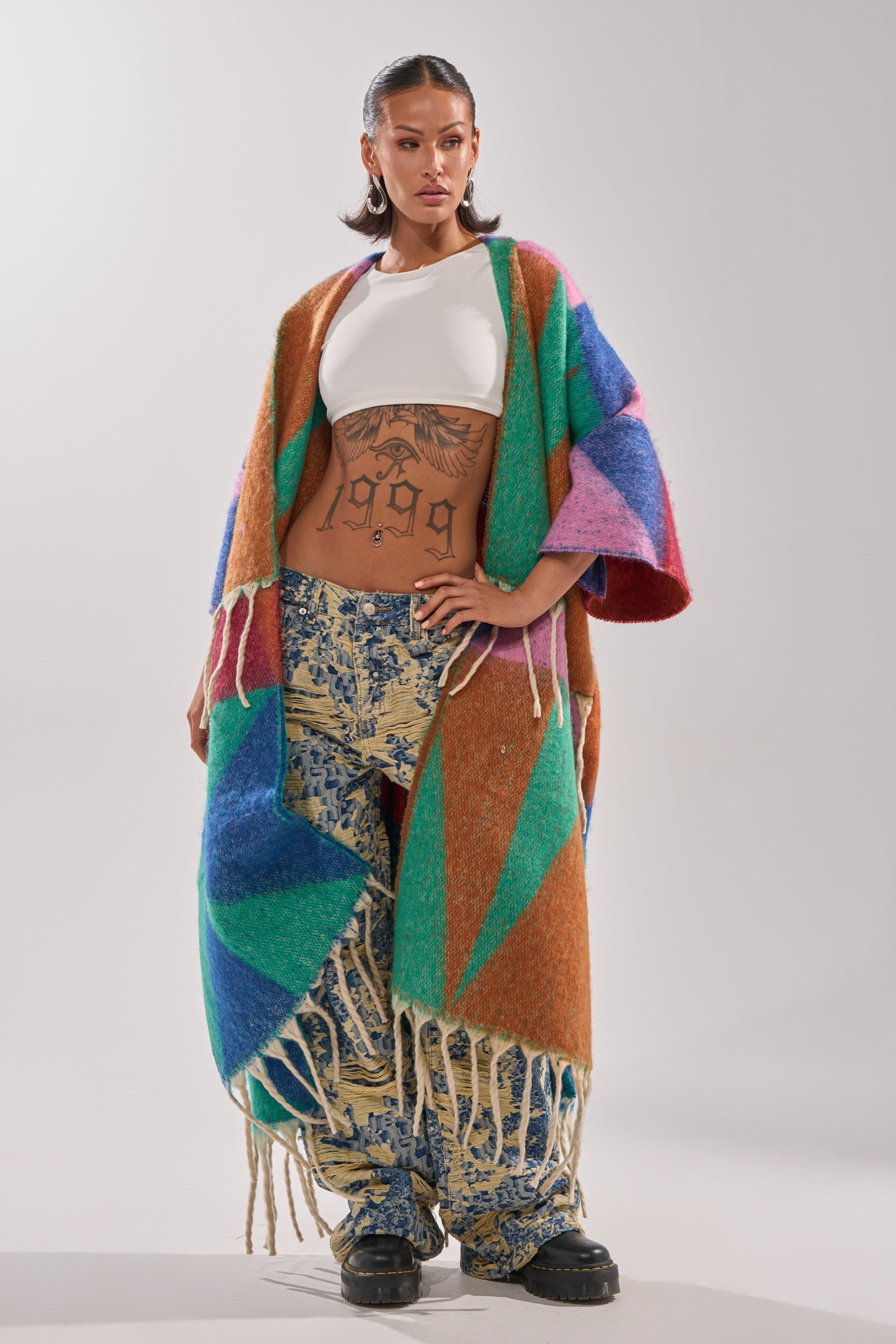 A woman stands confidently wearing the FEELING CRAZY OVERSIZED CARDIGAN as the highlight of her winter wardrobe, paired with a white crop top, blue patterned pants, black shoes, hoop earrings, and a visible "1999" tattoo across her torso.