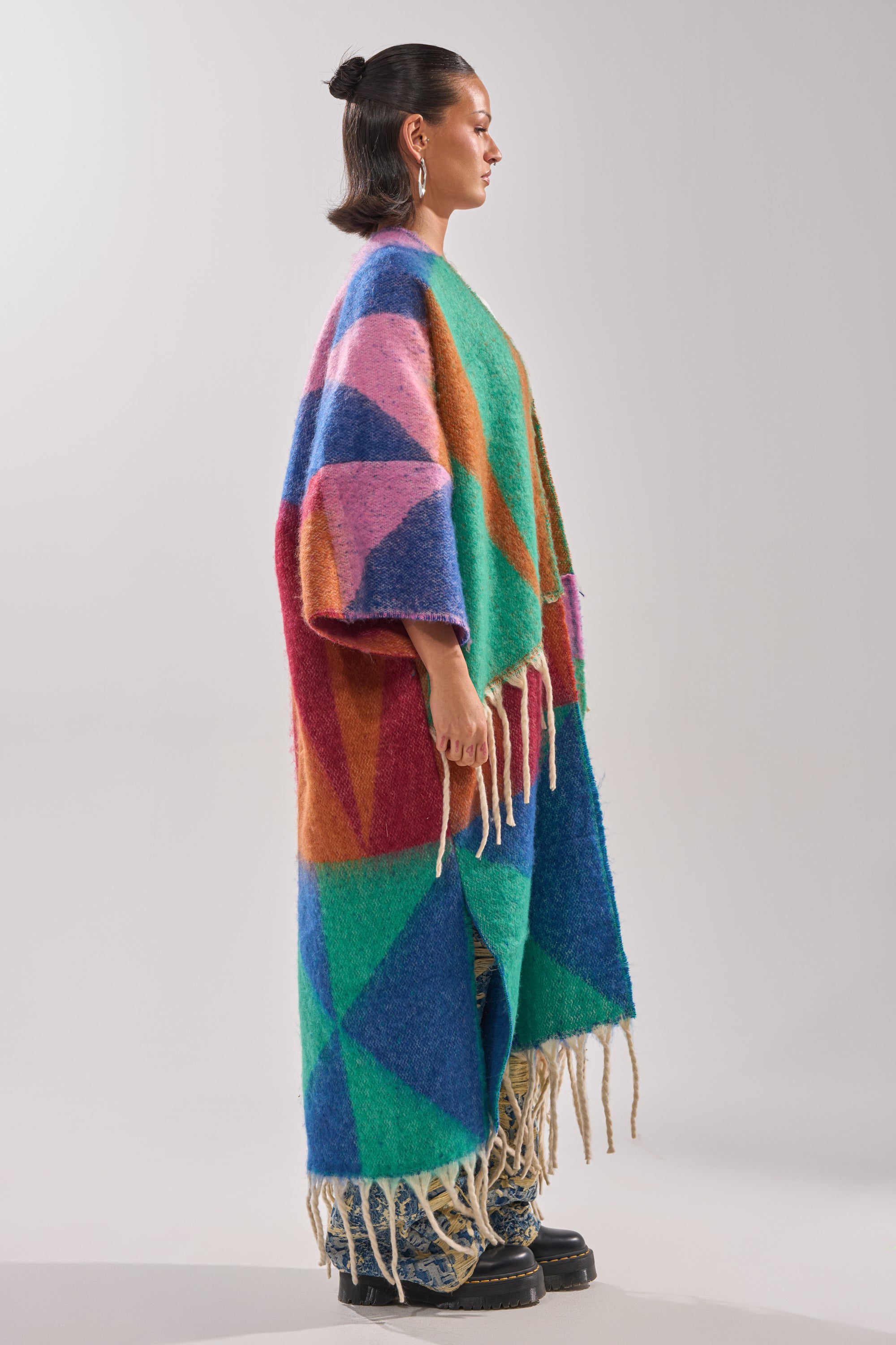 A person stands sideways against a plain light background wearing the FEELING CRAZY OVERSIZED CARDIGAN, a long colorful geometric sweater with fringe, styled with flared blue jeans and chunky black shoes.