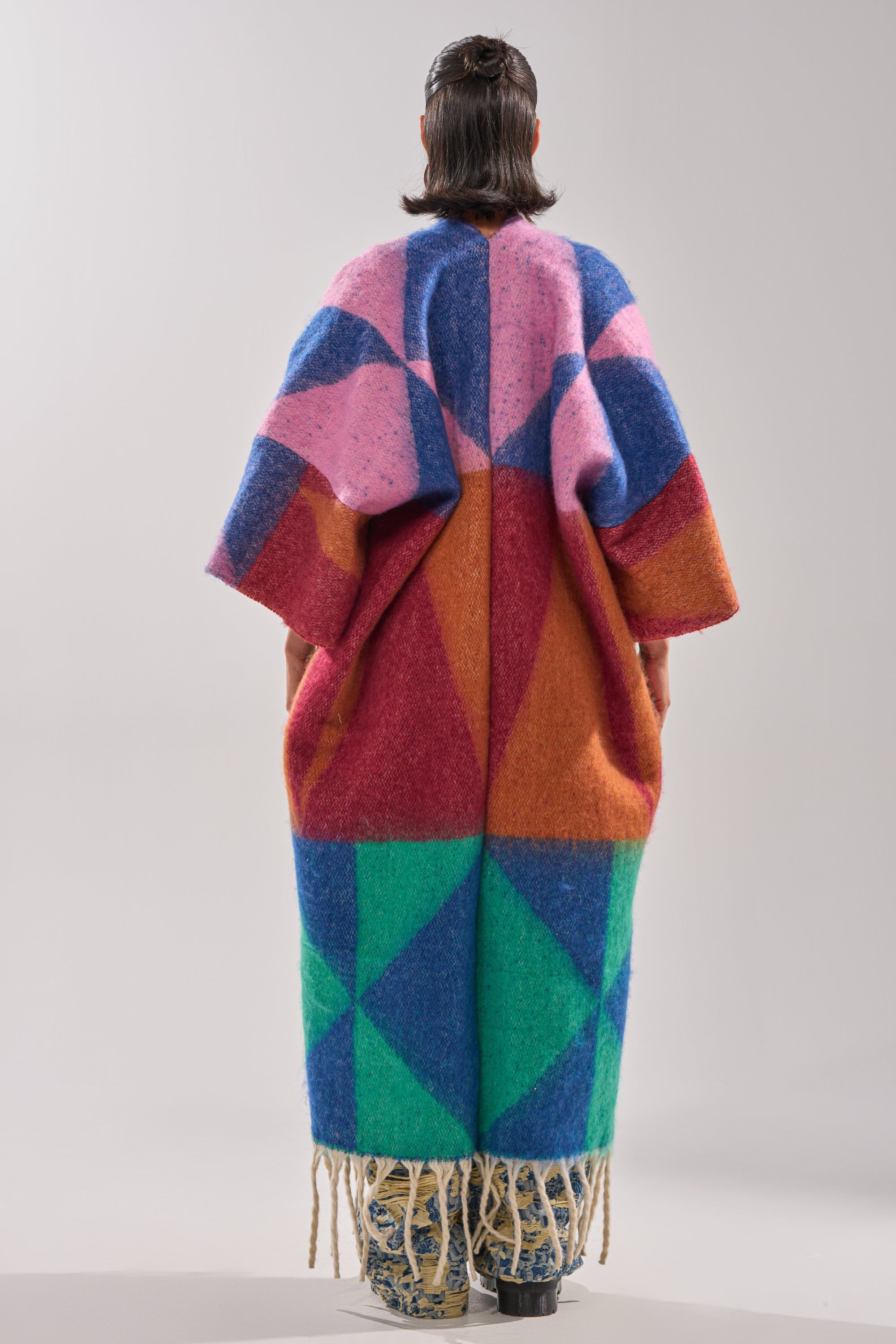 A person with shoulder-length hair stands facing away, wearing the FEELING CRAZY OVERSIZED CARDIGAN—a long, loose-fit sweater with bold geometric patterns in pink, blue, red, orange, and green plus fringe for extra flair.