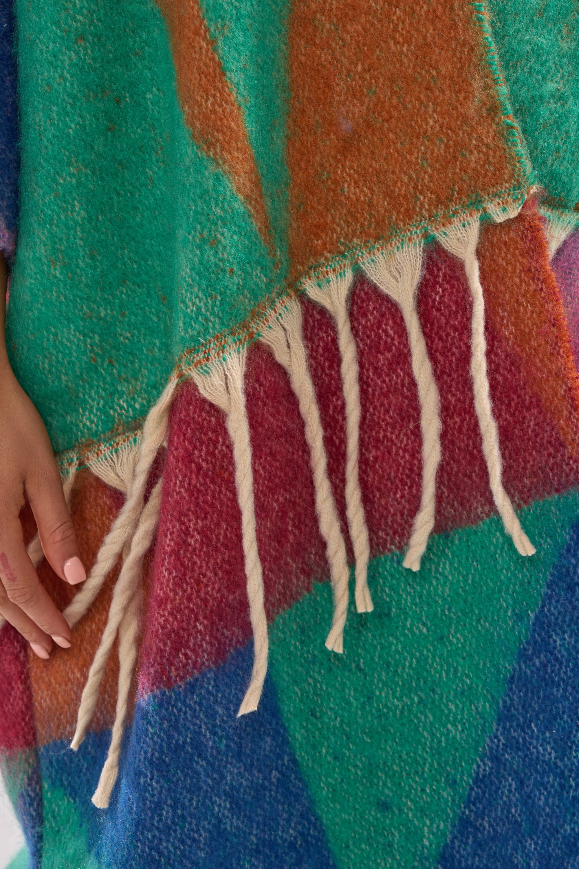 A close-up of the FEELING CRAZY OVERSIZED CARDIGAN, a multicolored knit with green, orange, red, and blue sections and fringe. A hand with pink nails gently touches it—perfect inspiration for your bold winter wardrobe.