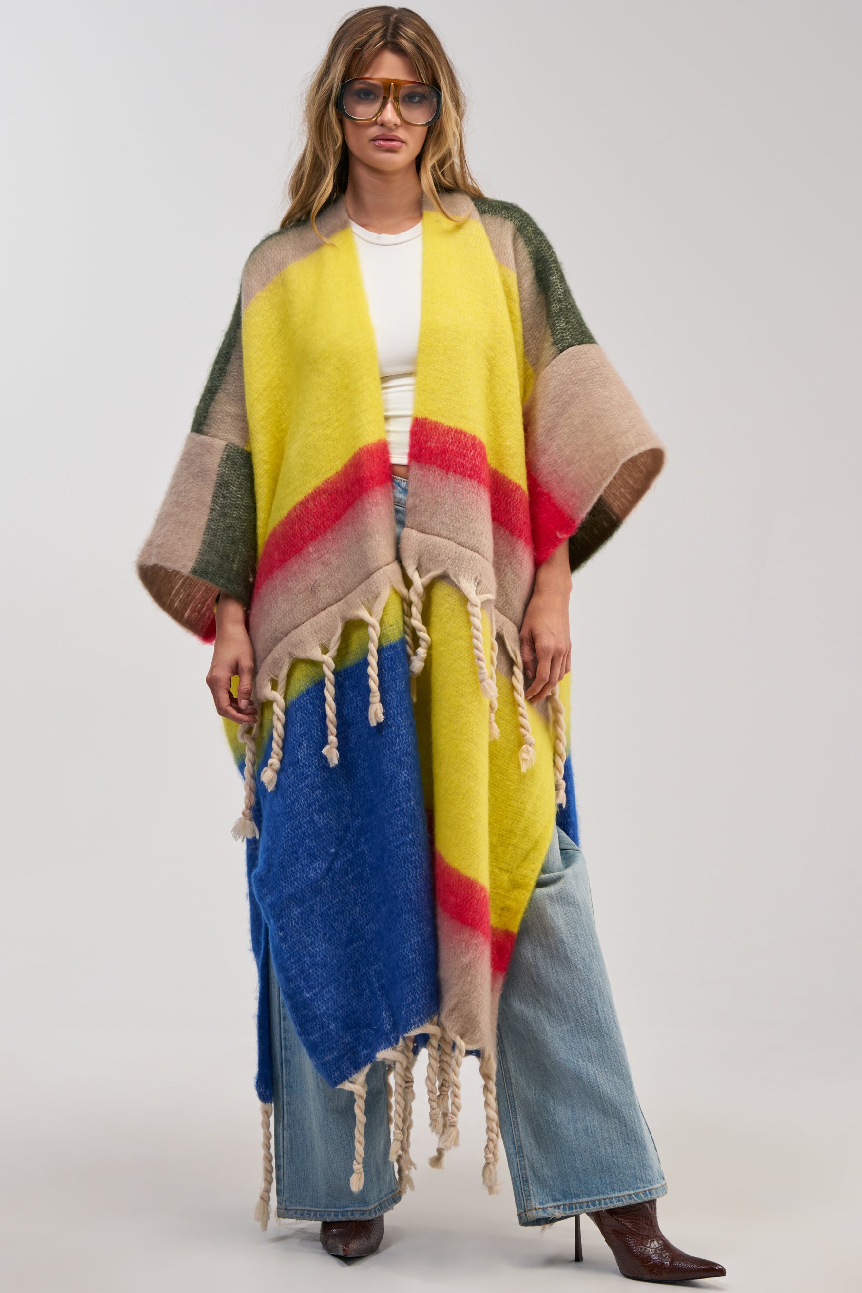 A person wearing the SUPER COZY OVERSIZED CARDIGAN IN YELLOW MULTI—a vibrant, colorful piece featuring yellow, red, blue, beige, and green sections reminiscent of an oversized fringe sweater—layered over a white top with wide-leg jeans, brown boots, and large glasses, standing against a plain background.