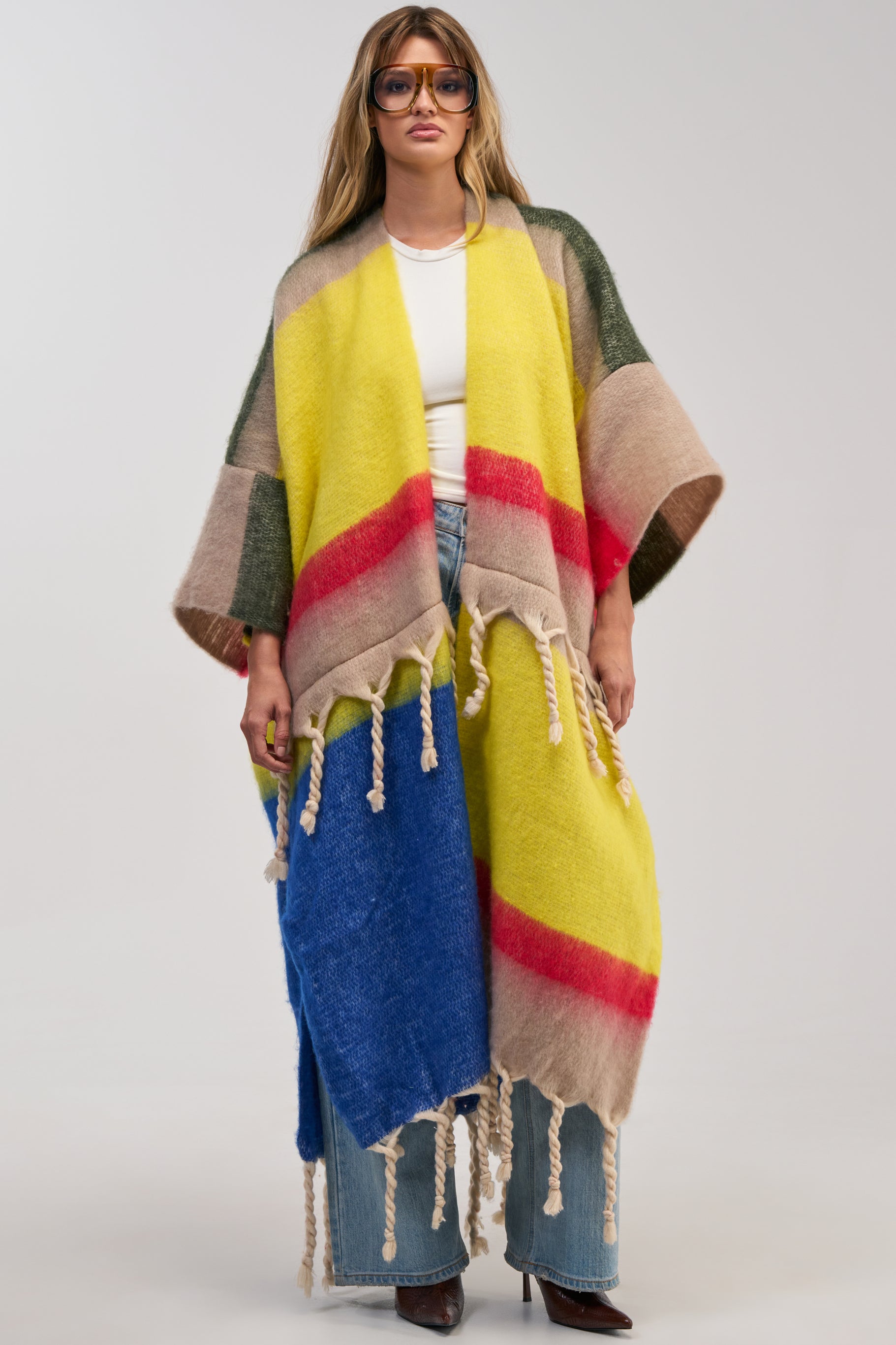 A woman wearing sunglasses, a white top, jeans, and the SUPER COZY OVERSIZED CARDIGAN IN YELLOW MULTI—featuring long, colorful fringes in yellow, red, beige, green, and blue—stands against a plain background.