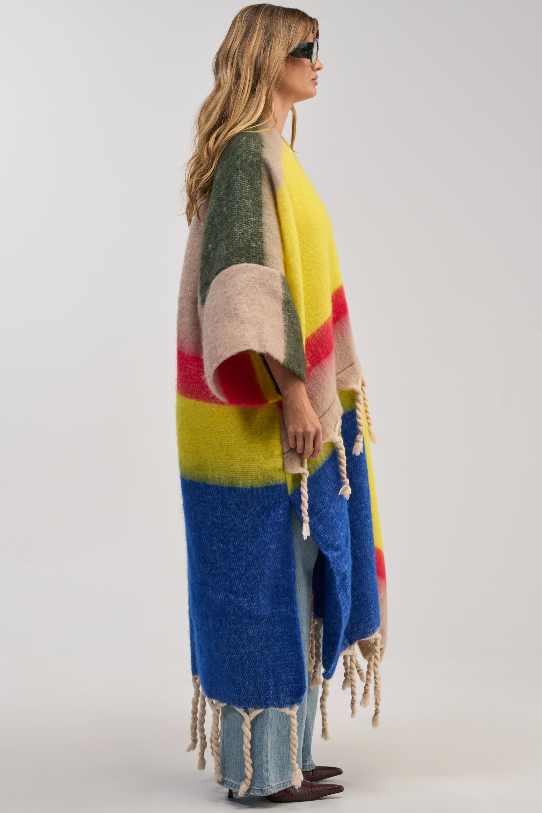 A woman in sunglasses wears the SUPER COZY OVERSIZED CARDIGAN IN YELLOW MULTI, styled as a loose, multicolored poncho with pink, yellow, green, and blue color blocks and thick fringed tassels, standing sideways against a plain background.