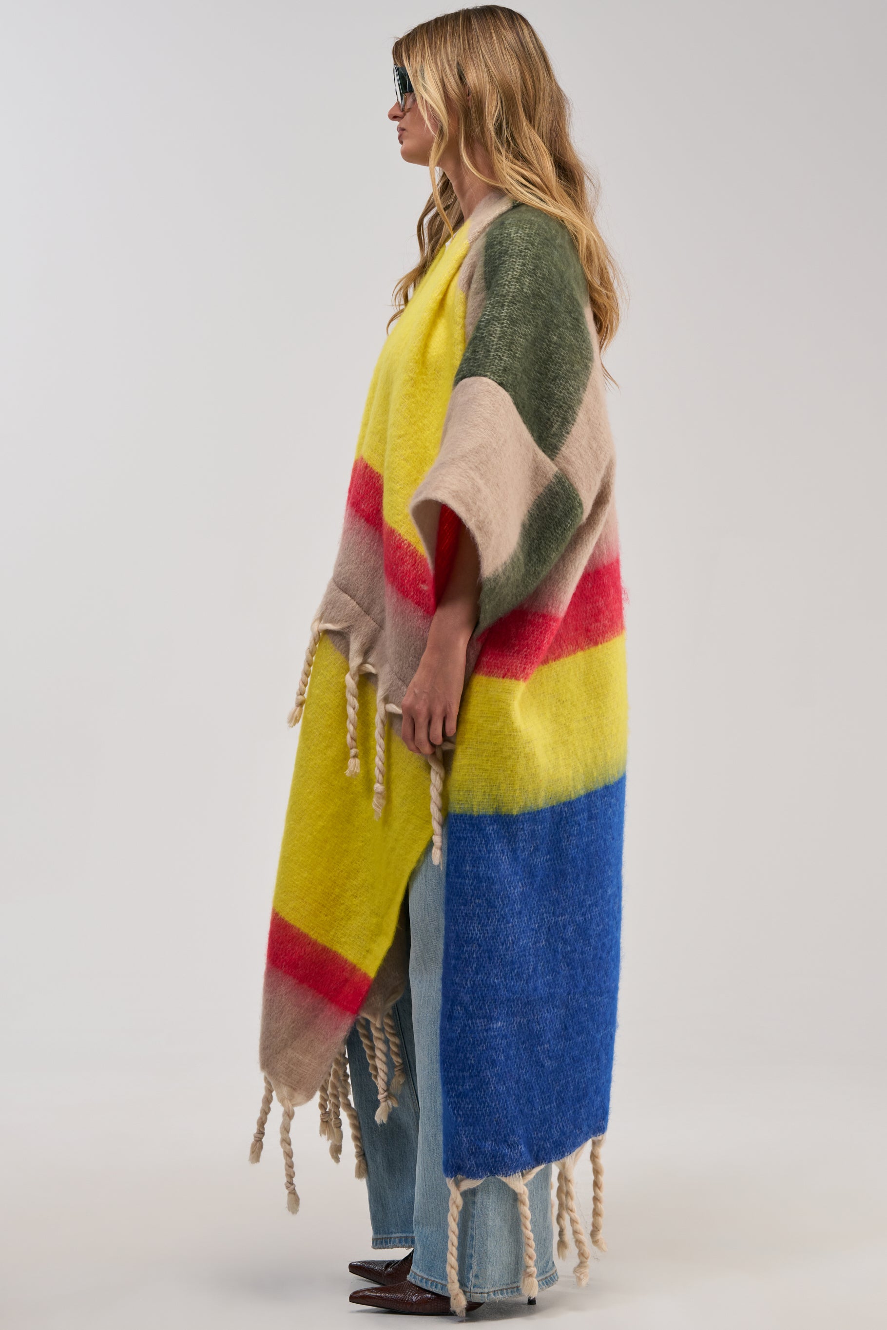 Wearing the SUPER COZY OVERSIZED CARDIGAN IN YELLOW MULTI—an eye-catching, long, colorful sweater featuring bold stripes and playful fringe details—a person pairs it with sunglasses, light blue jeans, and brown shoes while standing against a plain background.