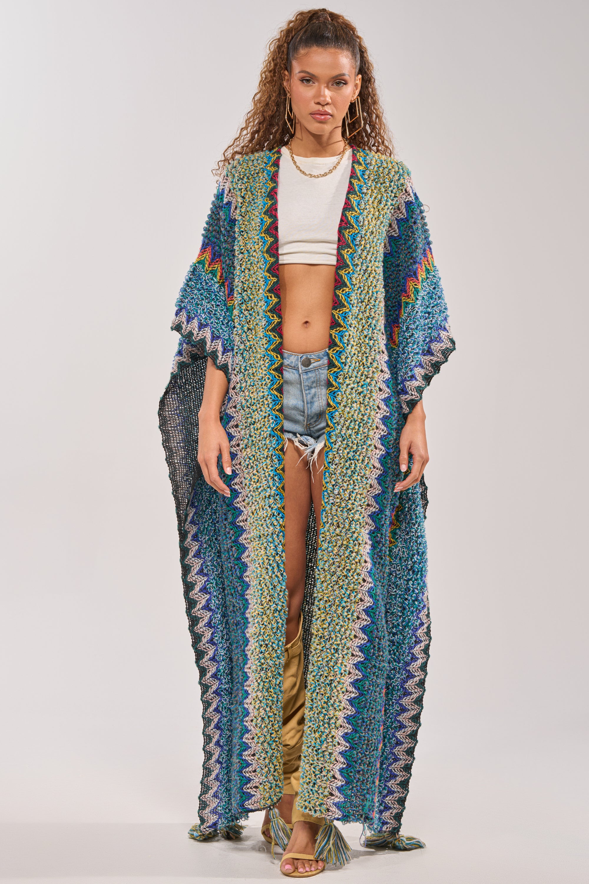A woman stands facing forward in the vibrant DESERT MOSAIC LONG CARDIGAN, perfect for festivals, styled over a white crop top and denim shorts. She accessorizes with curly hair, gold jewelry, and heeled sandals against a light background. 2