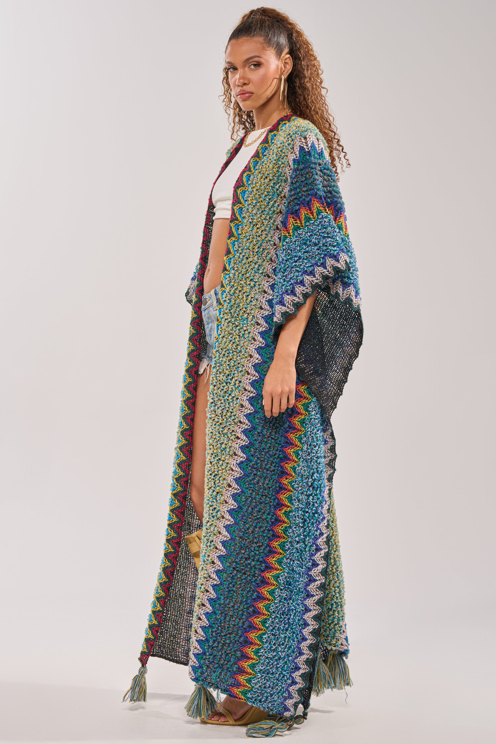 Wearing the DESERT MOSAIC LONG CARDIGAN with vibrant stripes and zigzag patterns, a woman stands sideways. Her curly hair is tied up, and she styles the knit duster with a white crop top and denim shorts for a festival-ready vibe. 3