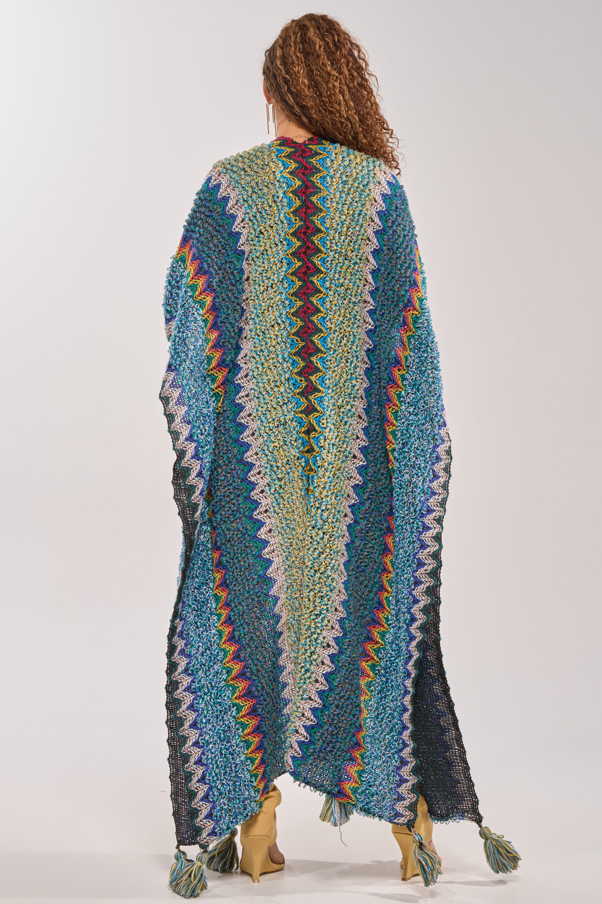 A person with curly hair faces away, wrapped in the DESERT MOSAIC LONG CARDIGAN featuring a colorful zigzag pattern and tasseled hem, set against a plain light background. 4