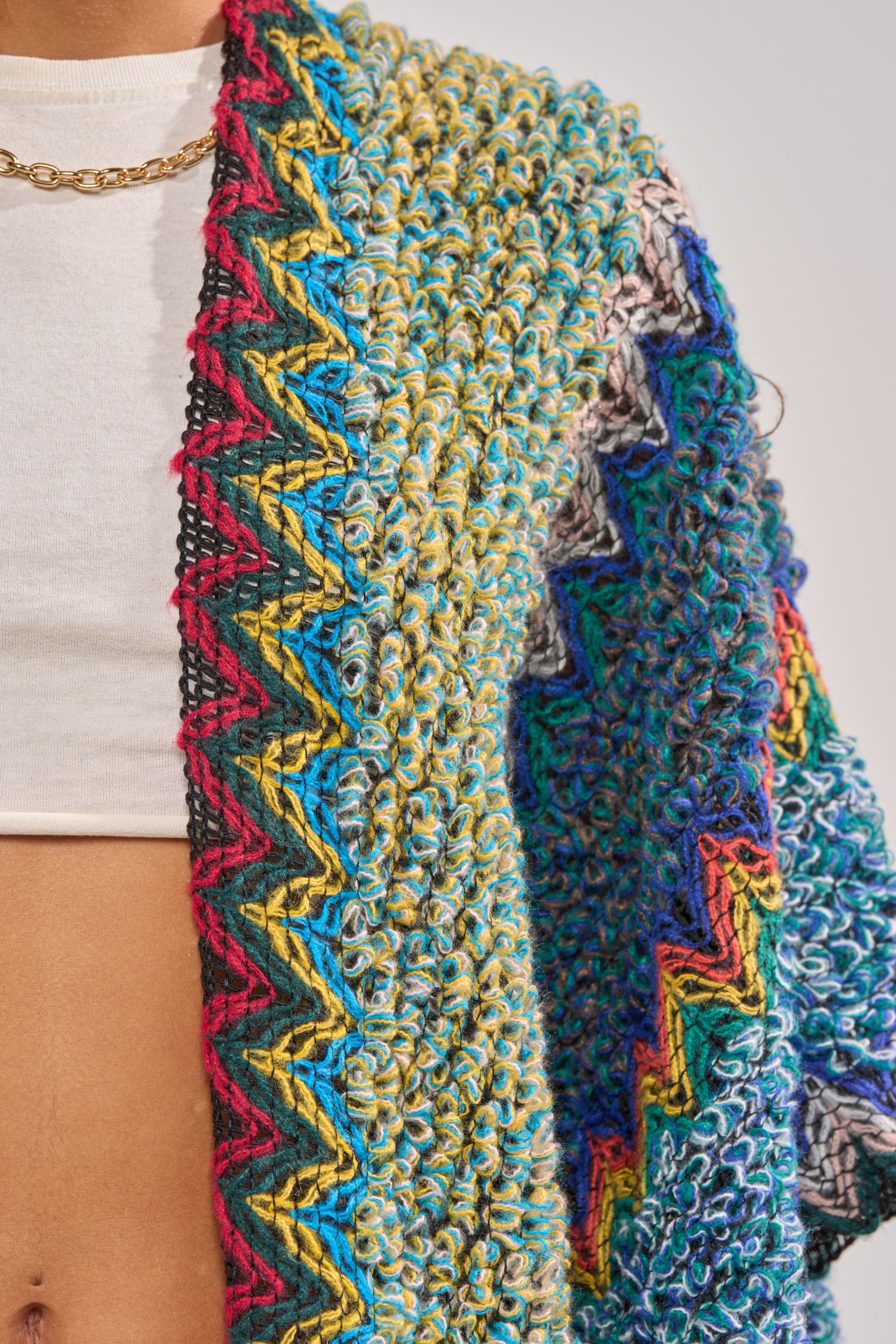 A close-up of a person wearing the DESERT MOSAIC LONG CARDIGAN, a colorful knit duster with bold zigzag patterns over a white crop top—ideal for festival looks. A gold chain necklace adorns their neck. 5