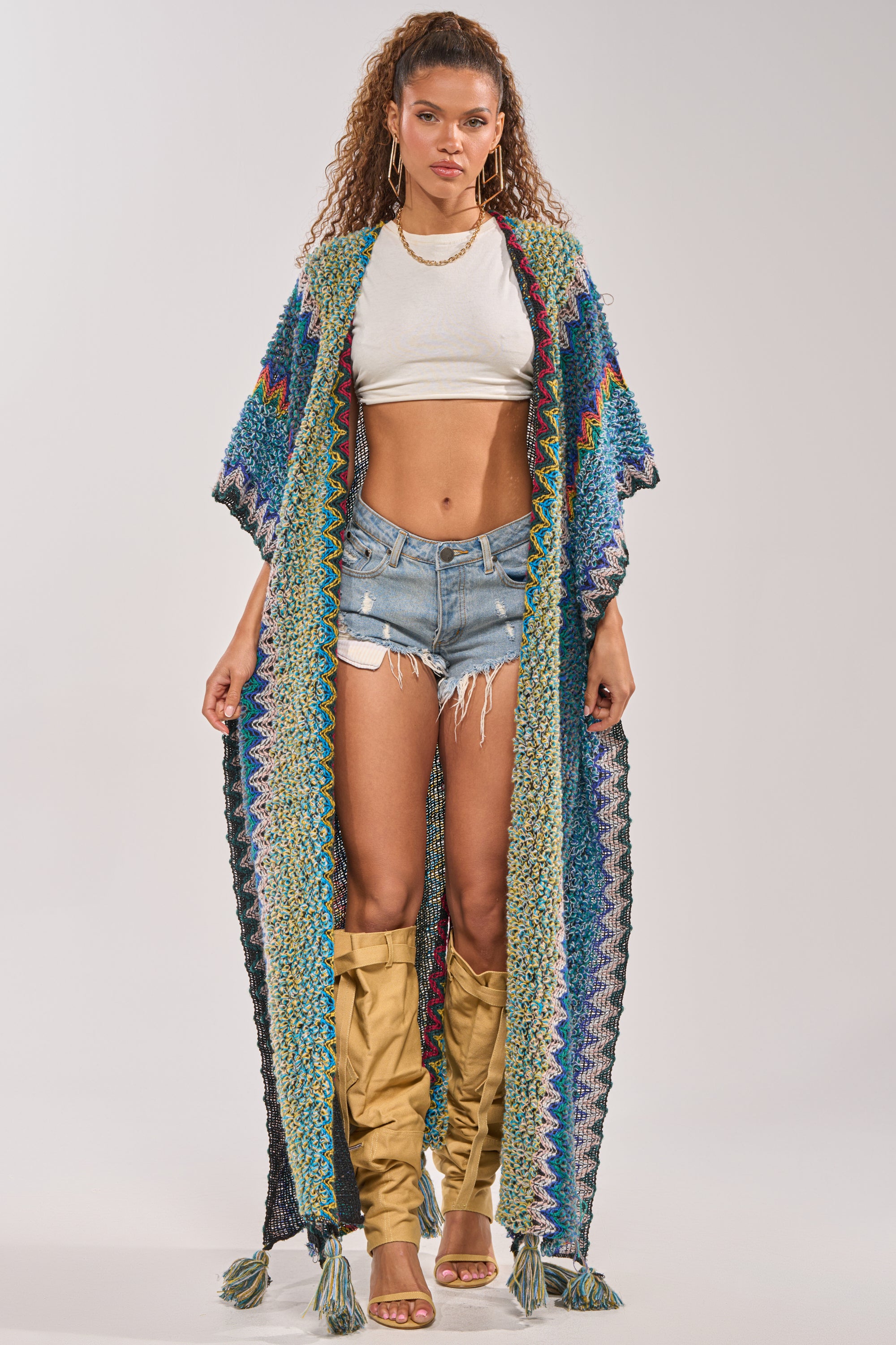 A woman with curly hair and a confident expression stands facing forward wearing the DESERT MOSAIC LONG CARDIGAN over a white crop top, distressed denim shorts, and gold accessories. 6