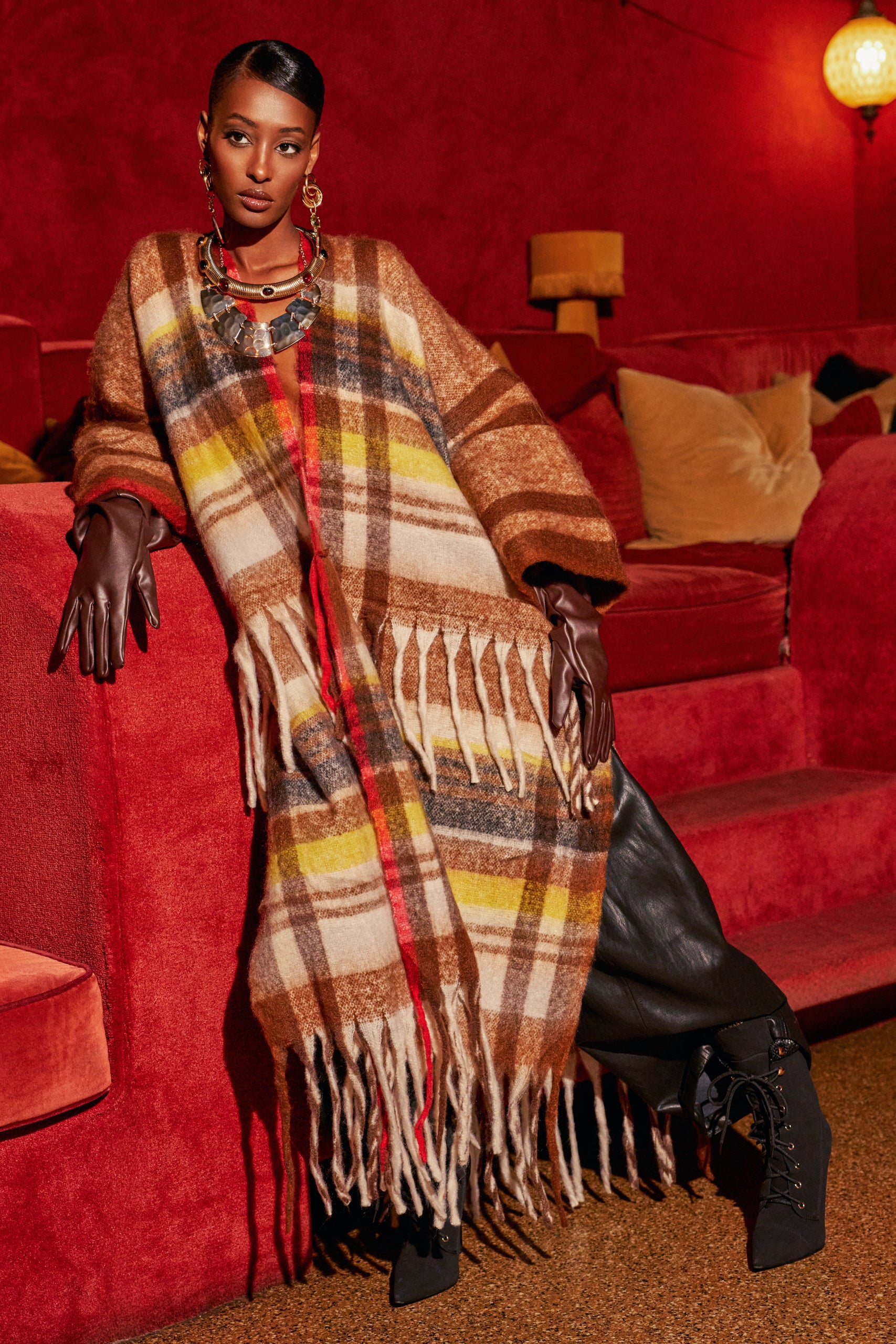 A person wears the AUTUMN HEIRLOOM CARDIGAN, a long, fringed, brown and yellow plaid piece, paired with dark brown leather gloves.