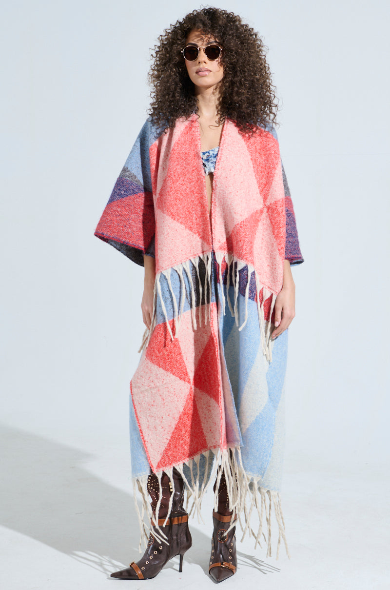 A woman with curly hair and sunglasses wears the BABY PLEASE LOVE CARDIGAN, a colorful geometric-patterned poncho featuring fringe tassels. She stands confidently in high-heeled boots against a plain background, emphasizing her bold, stylish look.