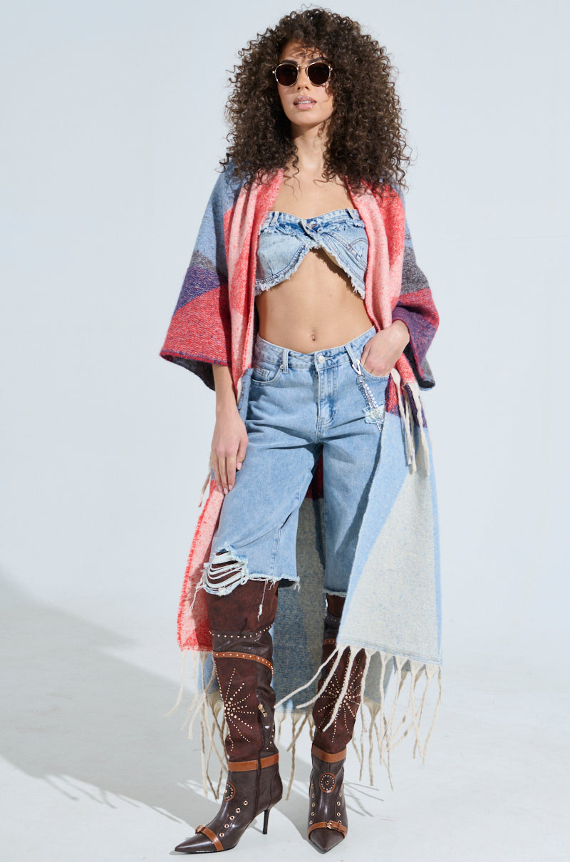 A woman with curly hair wears sunglasses, the BABY PLEASE LOVE CARDIGAN with geometric patterns, a ruffled denim crop top, distressed jeans, and tall brown heeled boots. She stands confidently against a plain background.