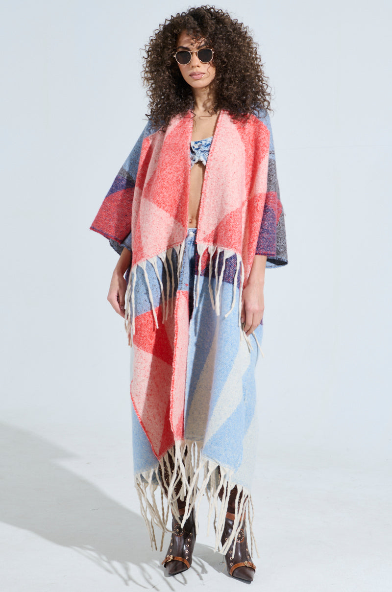 A person with curly hair wearing sunglasses, a BABY PLEASE LOVE CARDIGAN with colorful geometric patterns and fringe tassel detailing, blue jeans, and brown boots stands against a plain background.