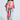 A person with curly hair wearing sunglasses, a BABY PLEASE LOVE CARDIGAN with colorful geometric patterns and fringe tassel detailing, blue jeans, and brown boots stands against a plain background.