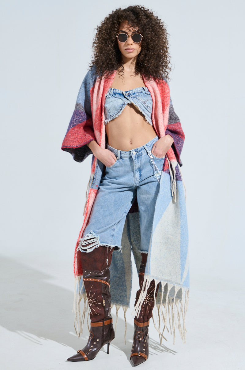 A person with curly hair confidently wears sunglasses, a colorful BABY PLEASE LOVE CARDIGAN with geometric patterns, a frayed denim top and jeans, brown studded high boots, and a tasseled scarf against a plain, light-colored background.