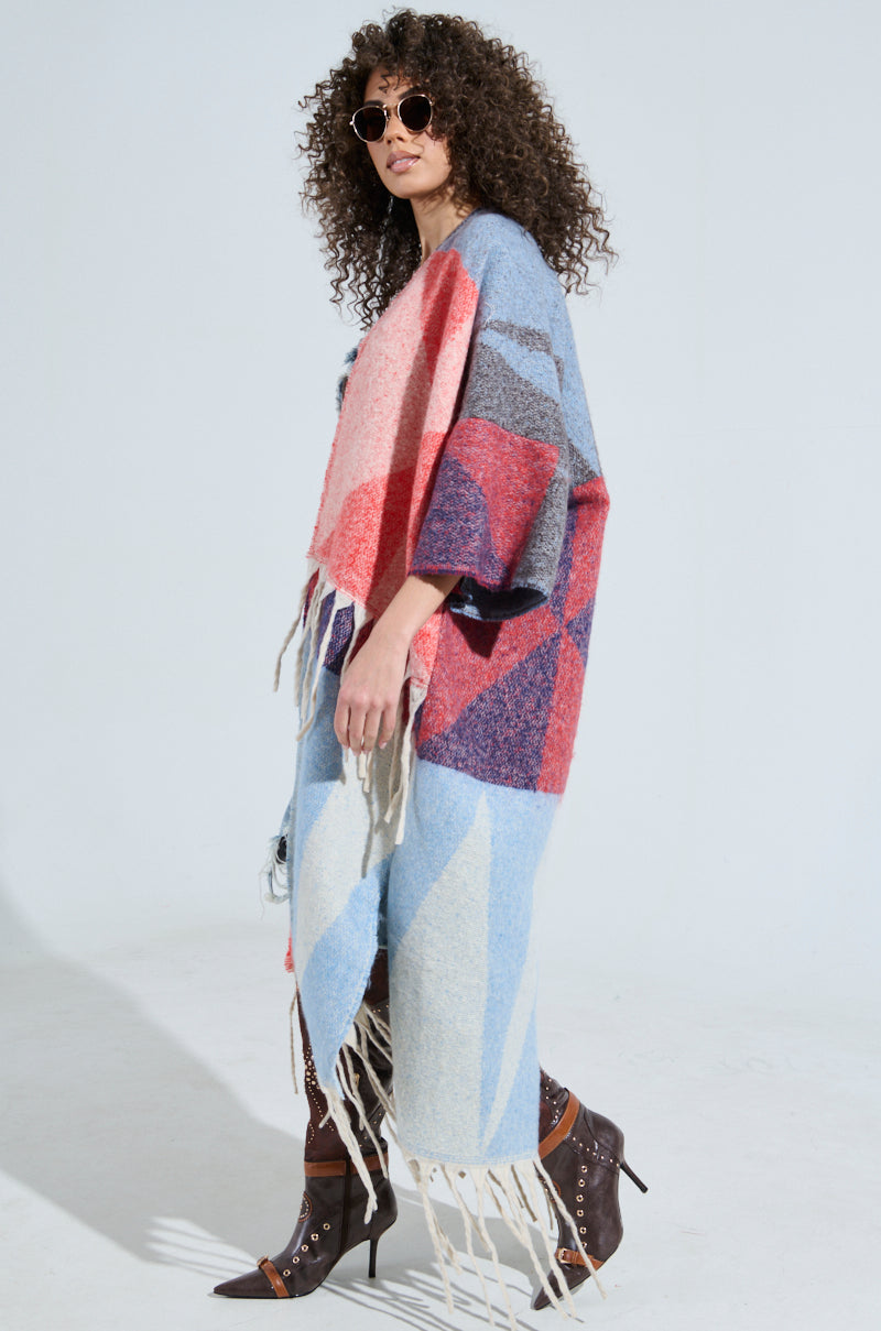 A person with curly hair walks sideways sporting sunglasses and studded knee-high boots while draped in the BABY PLEASE LOVE CARDIGAN, featuring a colorful geometric pattern with fringe tassel detailing in shades of blue, red, pink, and gray on a white background.