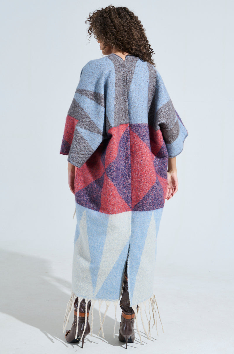 A person with curly hair wears the BABY PLEASE LOVE CARDIGAN in blue, gray, and red, adorned with fringe tassels and a geometric pattern. They're standing against a plain background facing away from the camera.