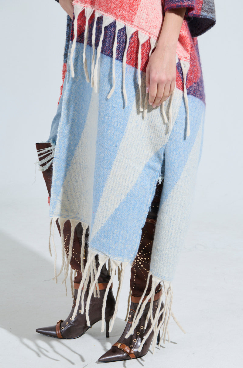 The BABY PLEASE LOVE CARDIGAN, adorned with geometric patterns in pink, blue, and red with fringe tassels, drapes over a person whose visible lower body is paired with pointed brown leather boots featuring decorative studs.
