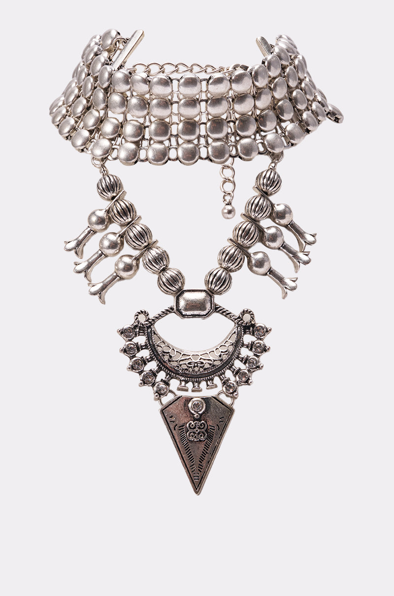 The IN MY VINTAGE ERA NECKLACE is a chunky silver-toned statement piece with a wide beaded collar, layered geometric and floral pendant, and dangling beads—perfect for adding festival flair.