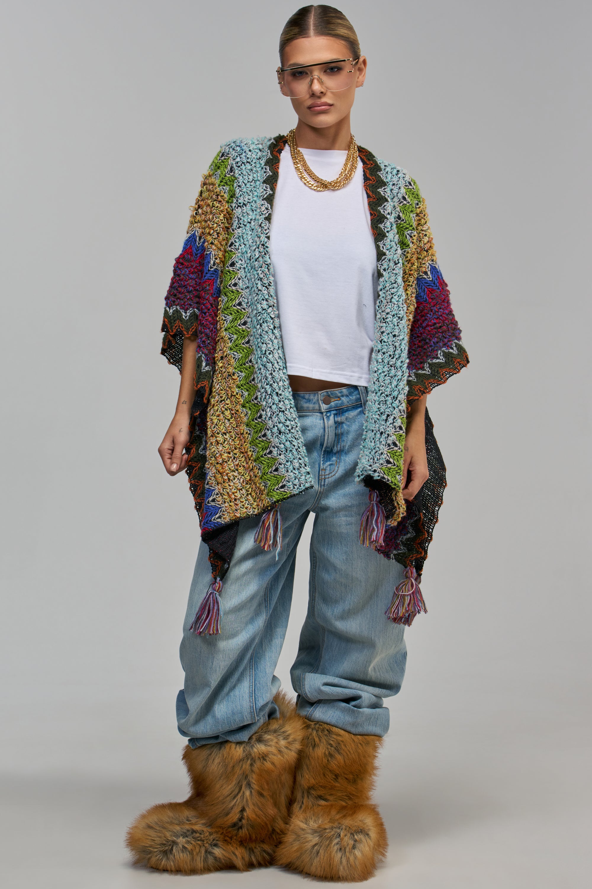 A person wears a SIMONE MULTI COLOR CARDIGAN IN GREEN MULTI, a colorful, open-front crochet knit cardigan with tassels, over a white shirt and baggy jeans, paired with shaggy faux fur boots for a bohemian streetwear look.