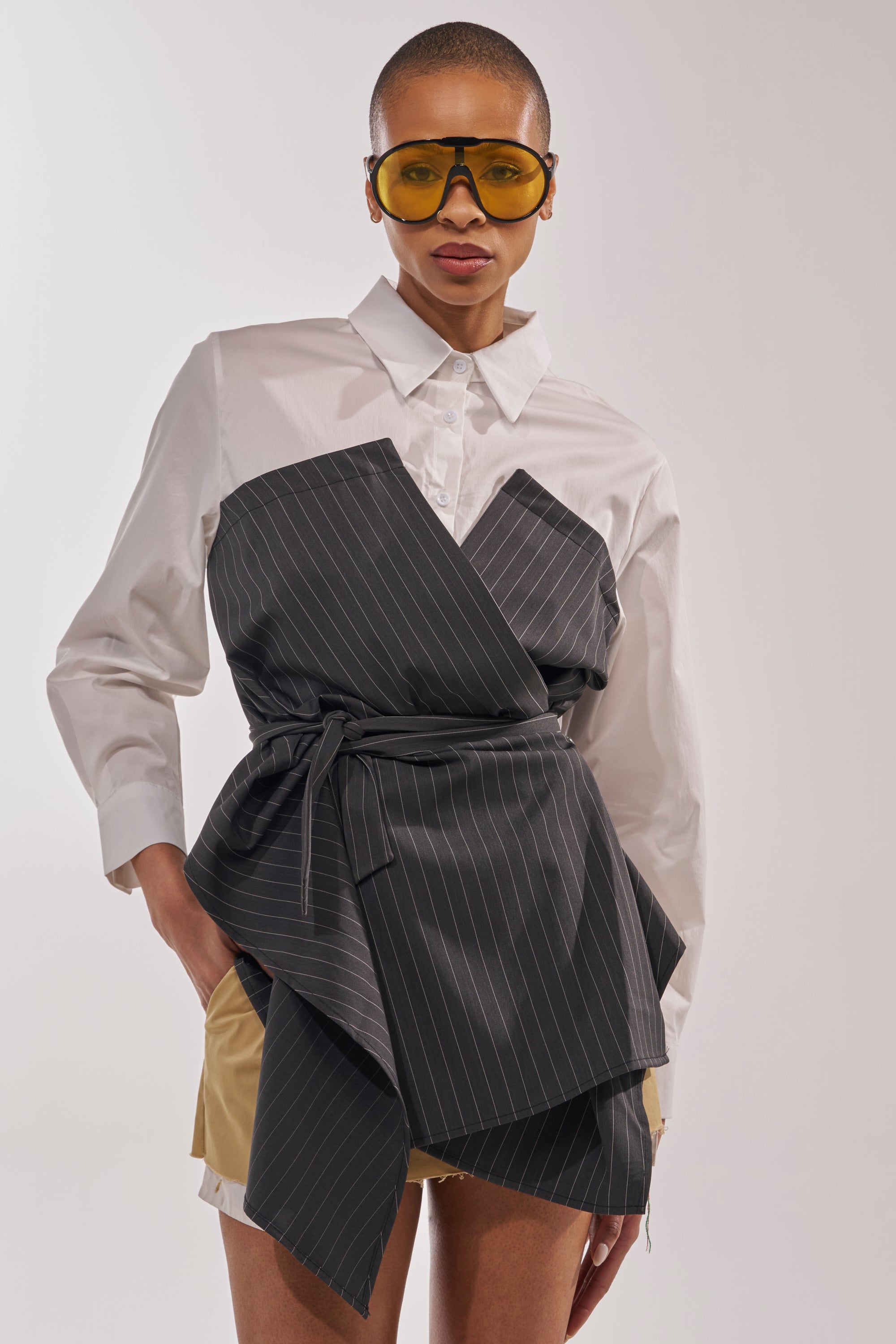 A stylish person confidently poses in yellow-tinted sunglasses, a white wrap-detail shirt layered over the BUSINESS ONLY BUTTON DOWN, and beige shorts, showcasing modern statement dressing against a plain background. 0