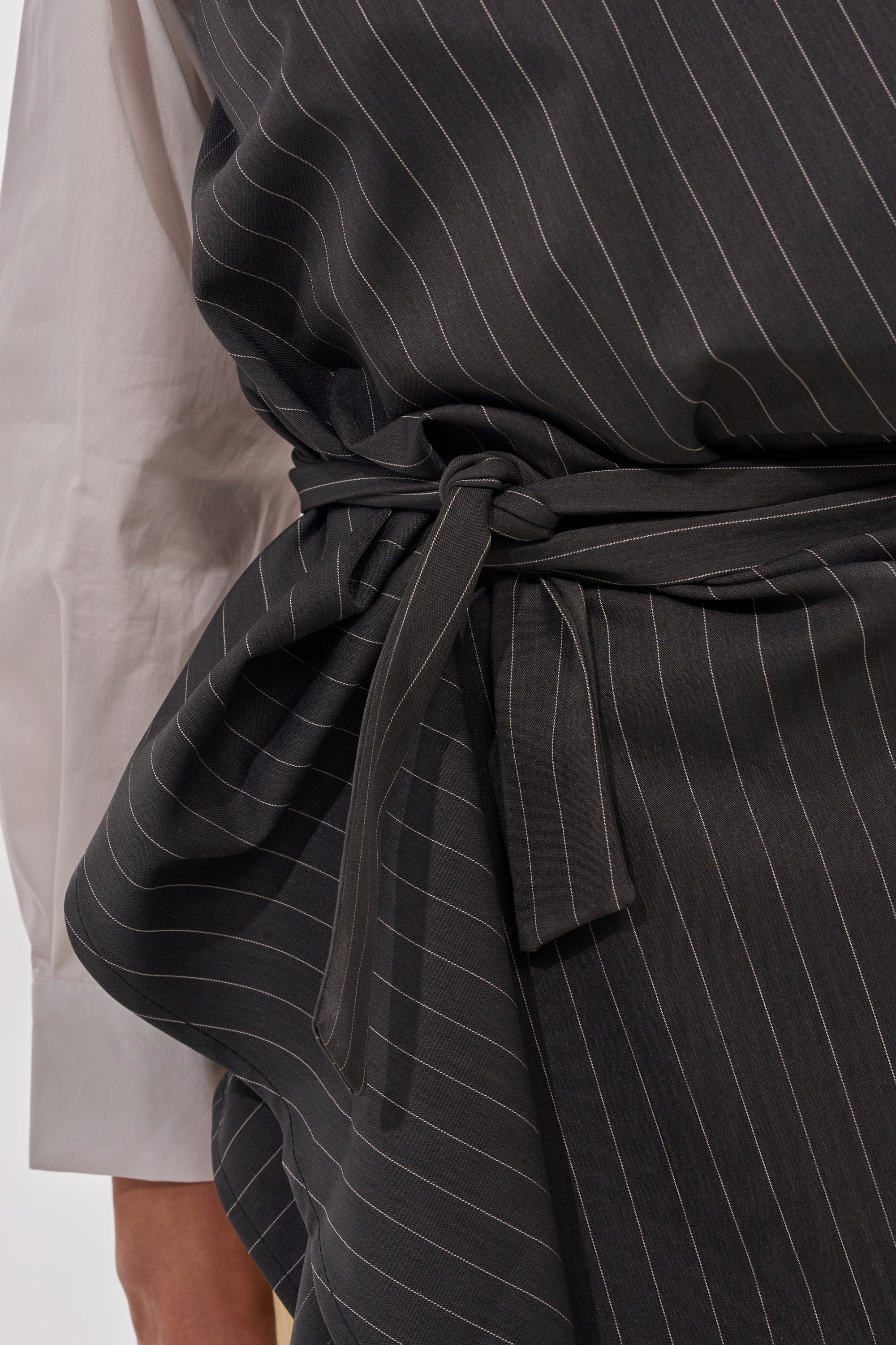 Close-up of a person wearing the BUSINESS ONLY BUTTON DOWN, a white shirt with dark gray pinstripes and a wrap-detail at the waist, secured by a matching belt. The textured fabric makes it perfect for office wear. 9