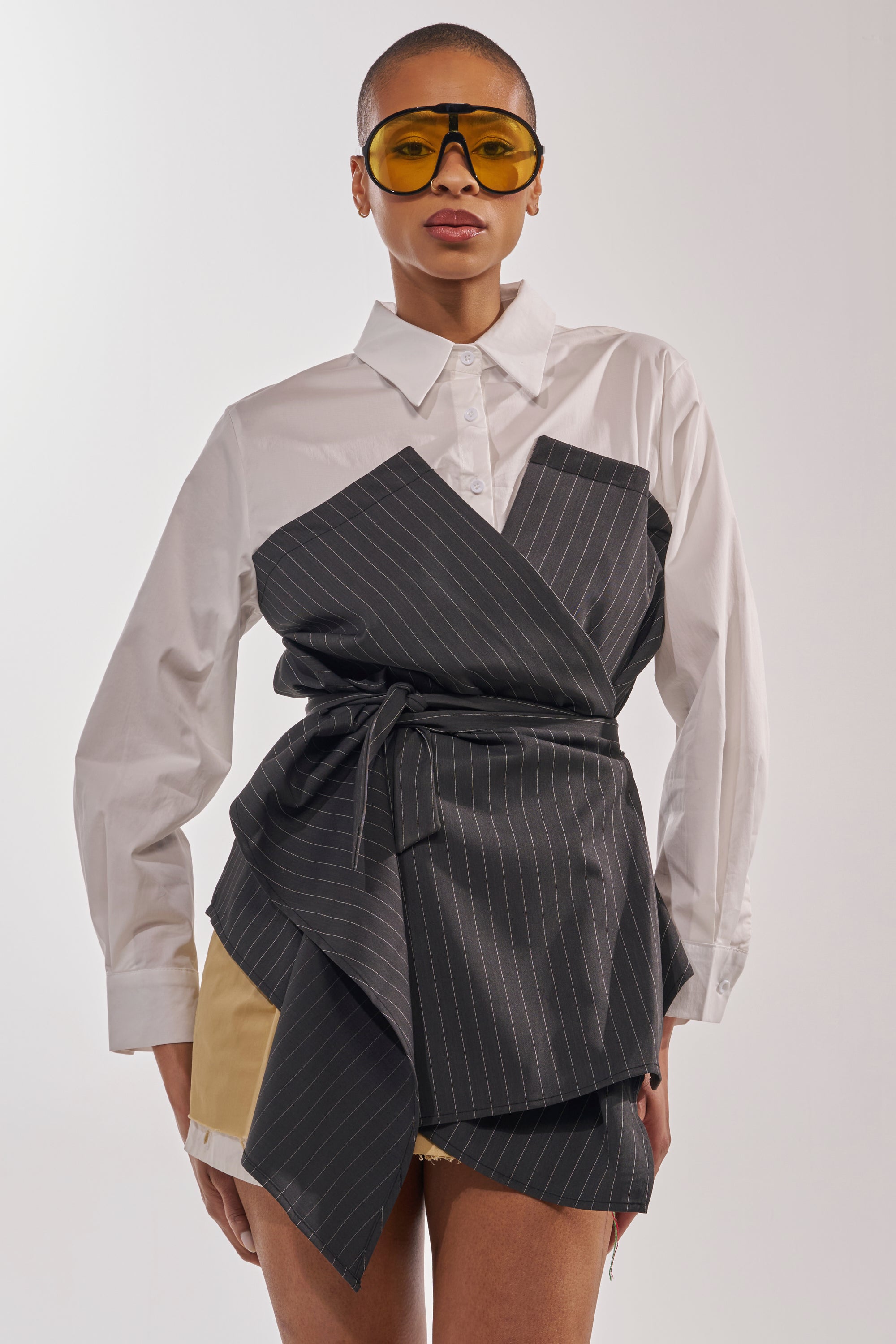 A person wears large yellow sunglasses and the BUSINESS ONLY BUTTON DOWN—a white pinstripe shirt styled as an asymmetrical wrap top with a tie—over tan shorts, standing against a plain light background. 1
