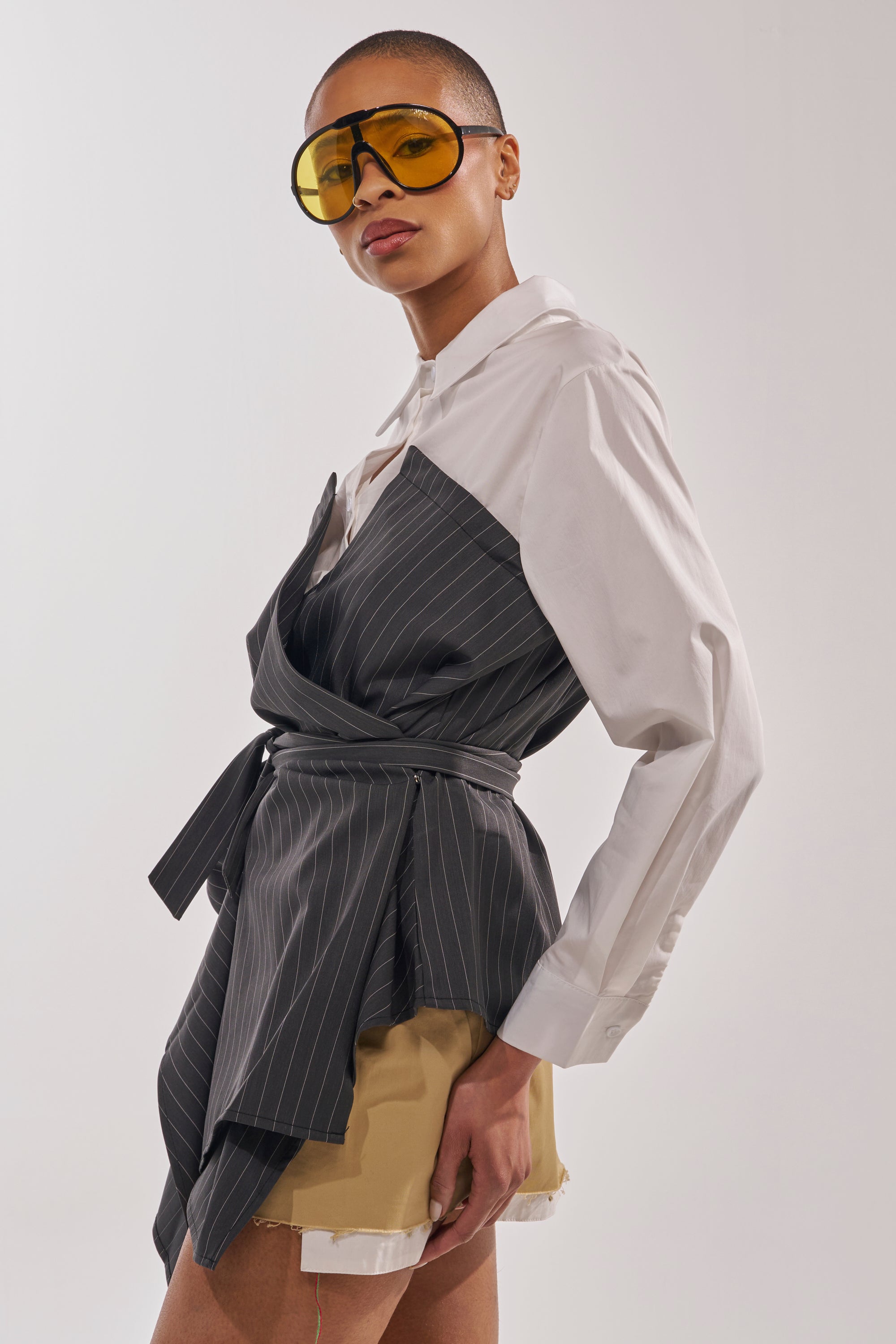 A stylish person with short hair wears yellow sunglasses, the BUSINESS ONLY BUTTON DOWN in white, a gray pinstripe wrap dress, and layered tan and white shorts, posing confidently against a plain background. 3