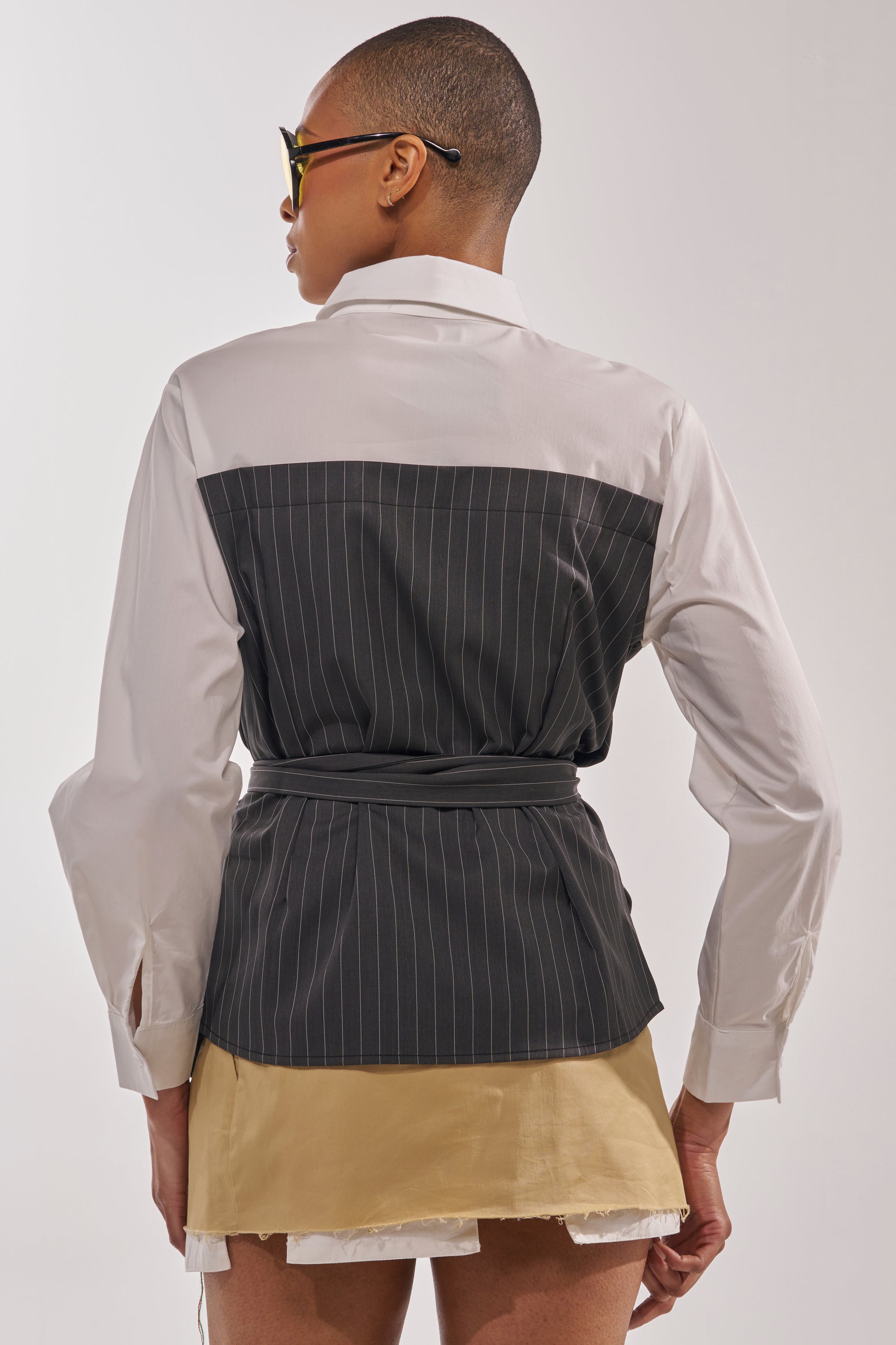 A person with short hair and sunglasses faces away, wearing the BUSINESS ONLY BUTTON DOWN—a pinstripe shirt with a belted waist and black back panel—paired with a beige mini skirt featuring a frayed white hem for chic office style. 5