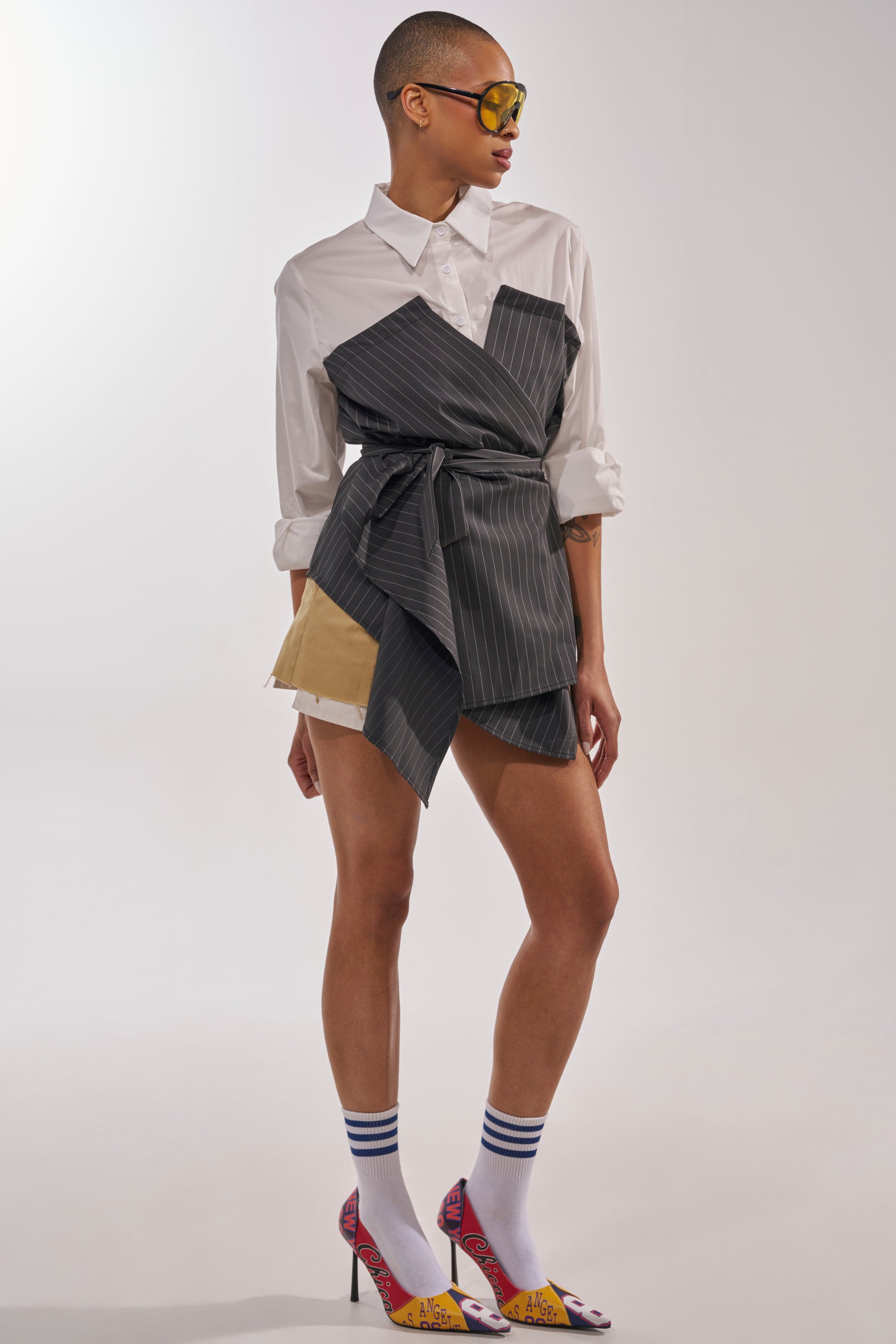 A person with a shaved head stands against a plain background, wearing the BUSINESS ONLY BUTTON DOWN styled as a wrap dress, yellow sunglasses, striped socks, and colorful high heels. 6